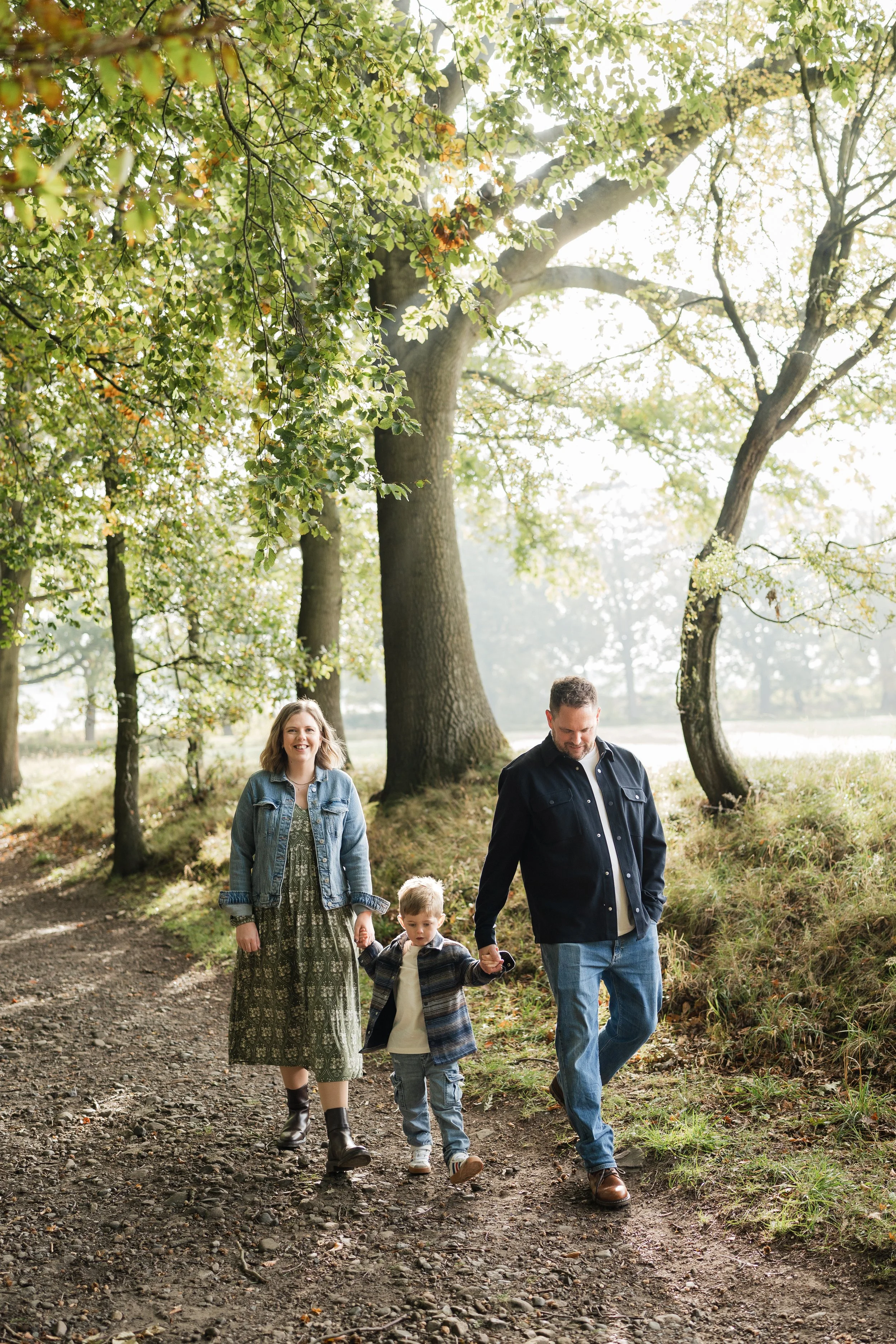 A family of three walking on a dirt trail through a wooded area with tall trees and green leaves during daytime. Newcastle family photos. 