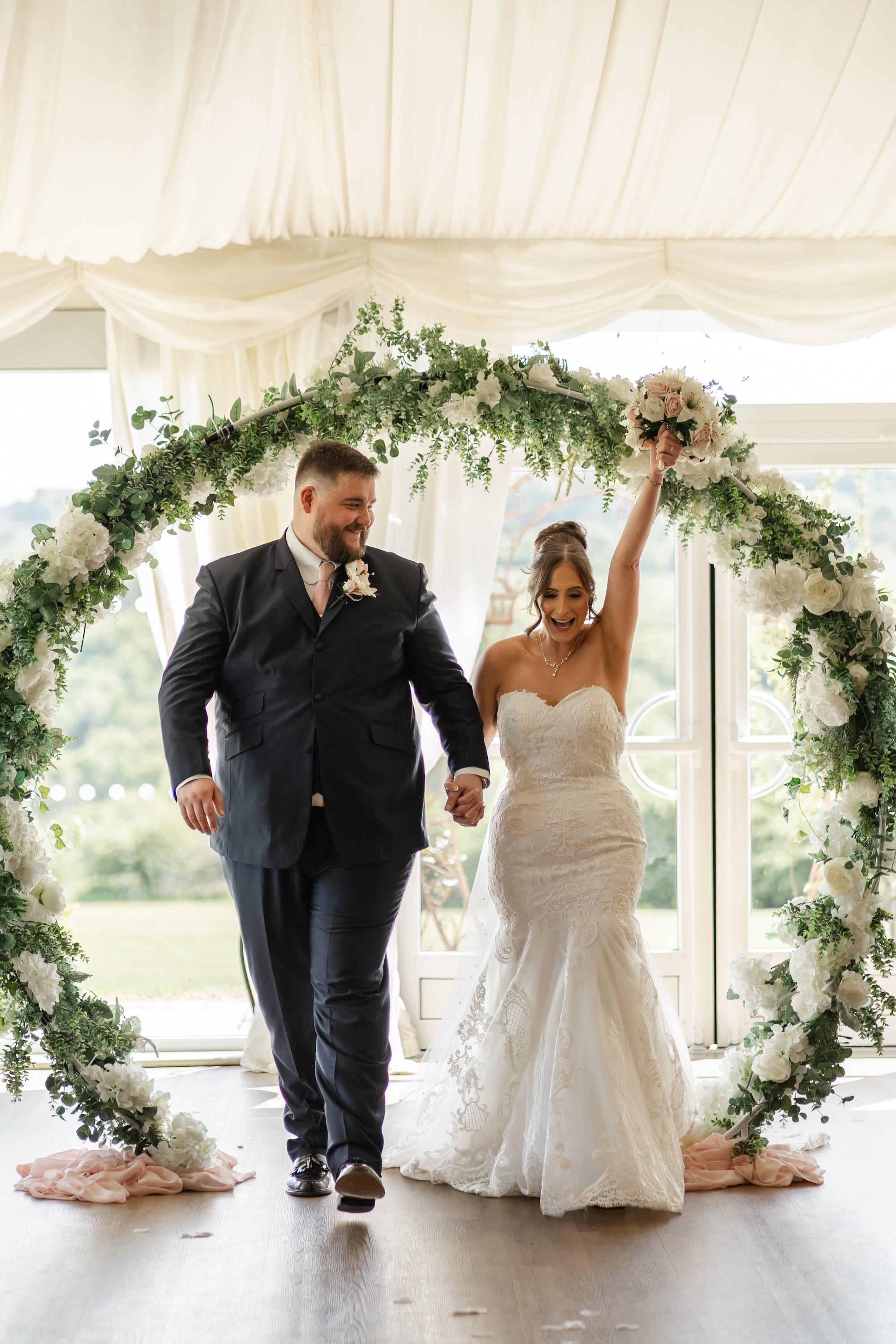 A newlywed couple is walking down the aisle under a floral arch, with the bride holding a bouquet and smiling, while the groom holds her hand and looks at her.