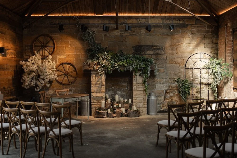 Interior of a rustic wedding or event space with wooden chairs arranged facing a fireplace decorated with greenery, candles, and flowers, brick walls, and soft lighting. Horton Grange barn. 