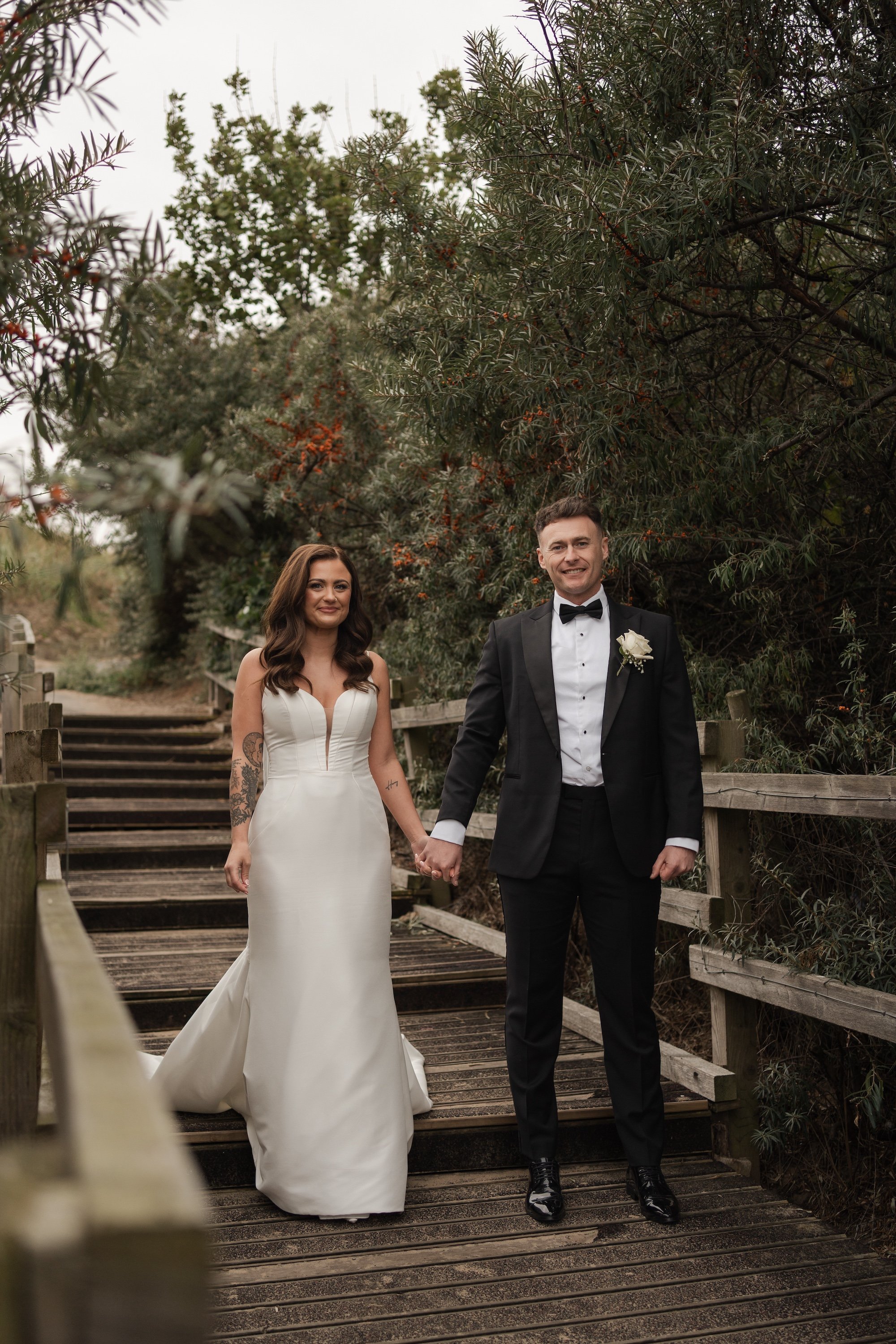 A bride and groom holding hands on a wooden pathway surrounded by green trees during their wedding. Tynemouth wedding photographer.