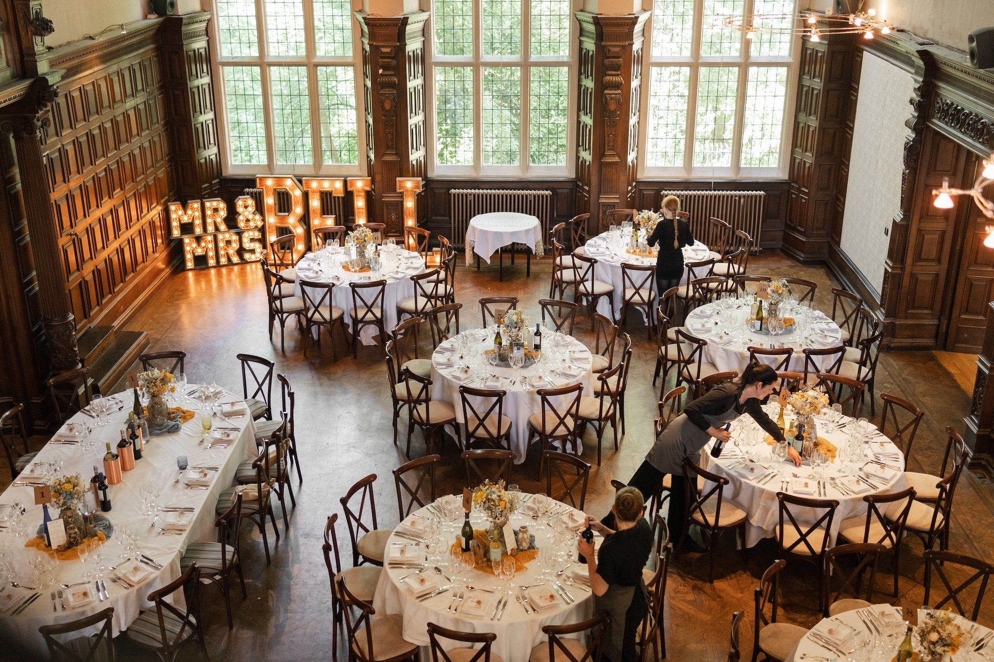 Wedding reception setup in an elegant wood-paneled banquet hall with round tables covered in white tablecloths, decorated with floral centerpieces, wine bottles, and table settings. Large windows let in natural light. In the background, illuminated m