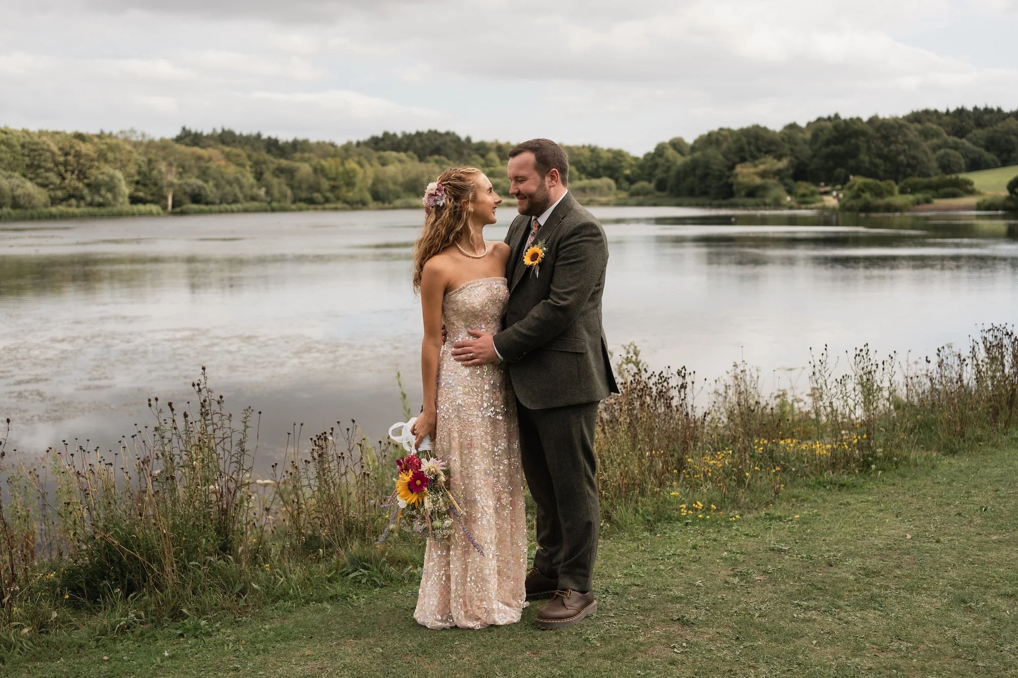 A bride and groom stand close together near a lake, smiling at each other on their wedding day. The bride wears a floral hairpiece and a strapless, beaded gown, holding a bouquet of colorful flowers. The groom wears a dark gray suit with a sunflower 