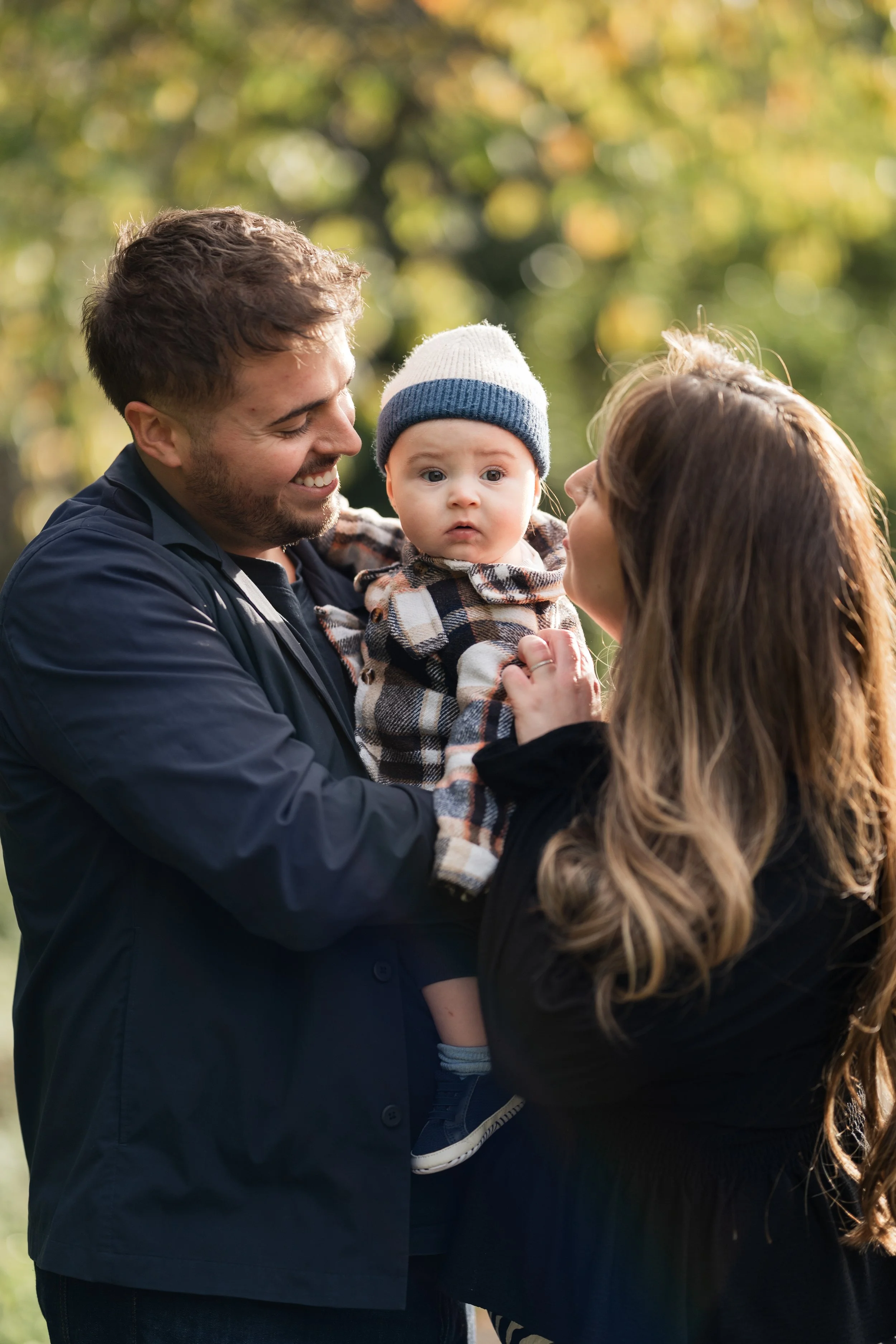 A happy man and woman holding a young child outdoors in a park with trees and sunlight in the background. Jesmond family photos. 