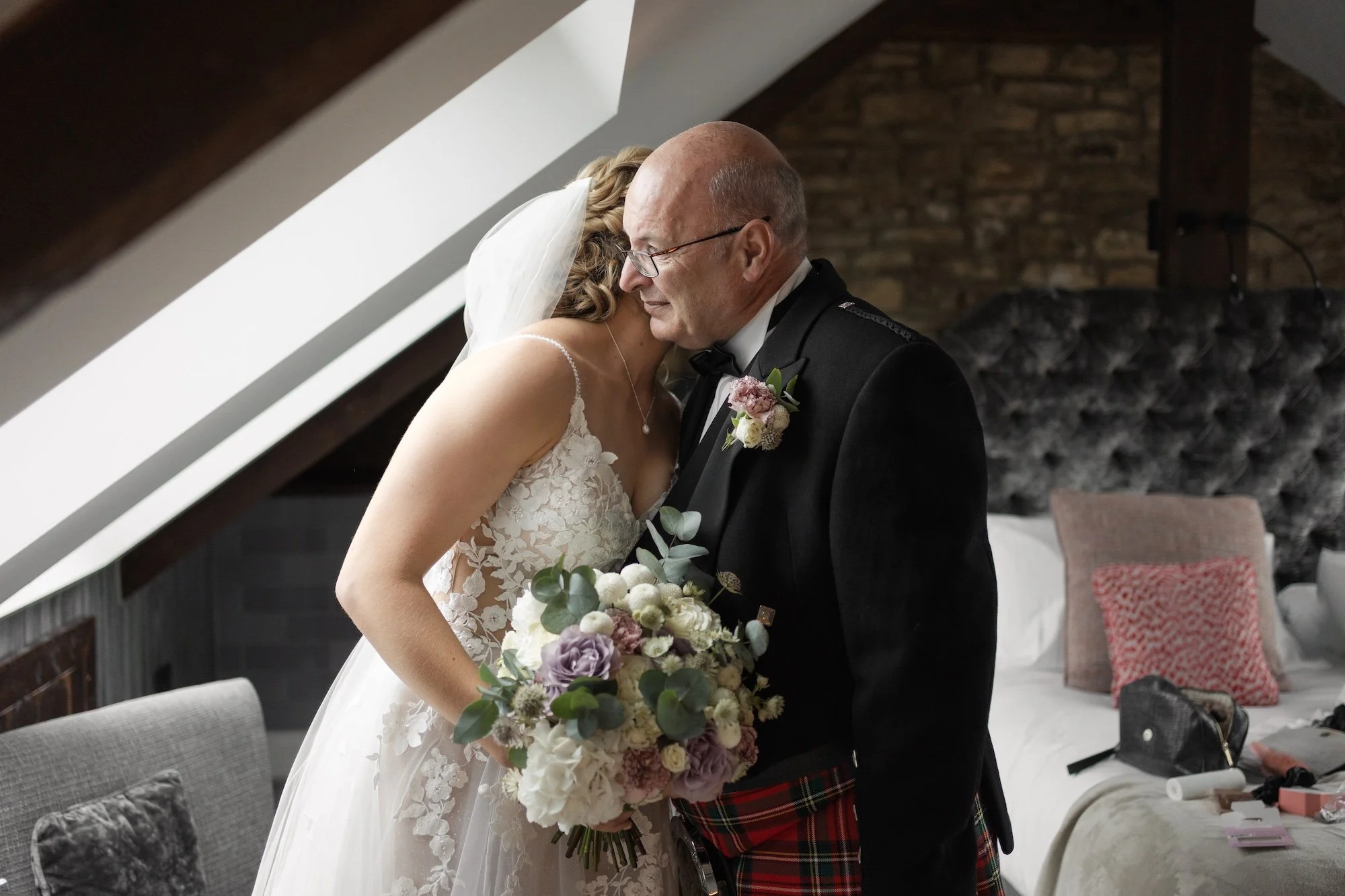A bride and an older man, dressed in formal wedding attire including a tuxedo with tartan kilt, sharing an emotional moment indoors with a brick wall and a bed in the background.