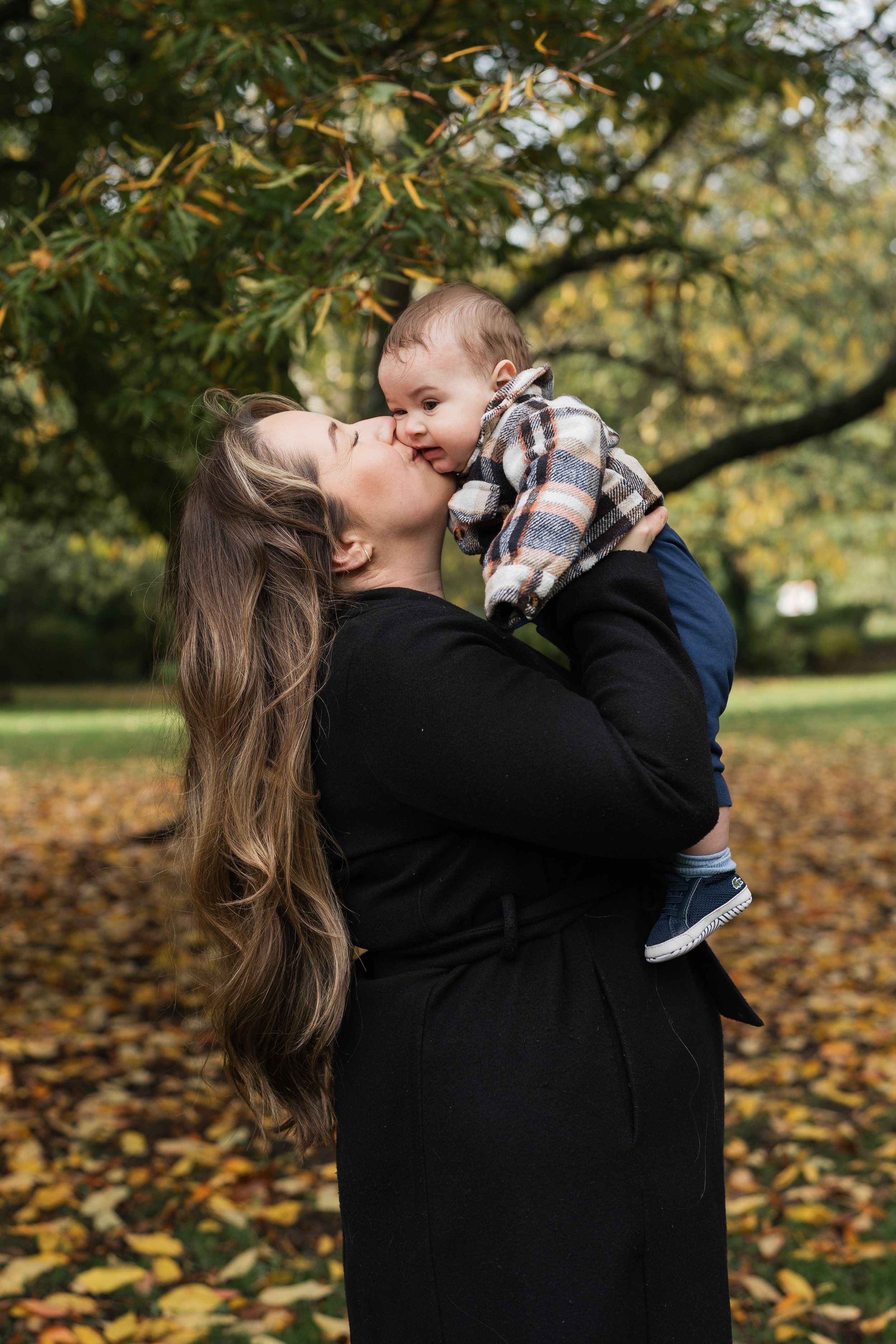 A woman with long brown hair holding a baby boy, giving him a kiss on the nose outdoors during autumn with fallen leaves and trees in the background.