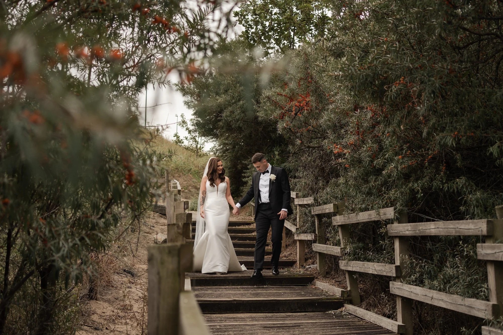 Bride and groom holding hands while walking down a wooden pathway surrounded by trees. Longsands, Tynemouth. 