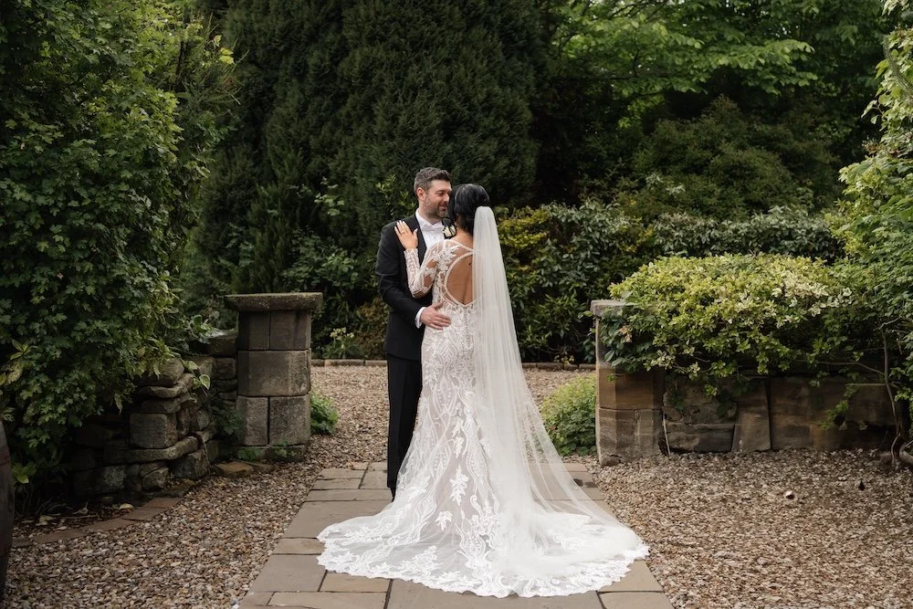 A bride and groom embracing in a garden, with the bride wearing a white lace gown and veil, and the groom in a black tuxedo.