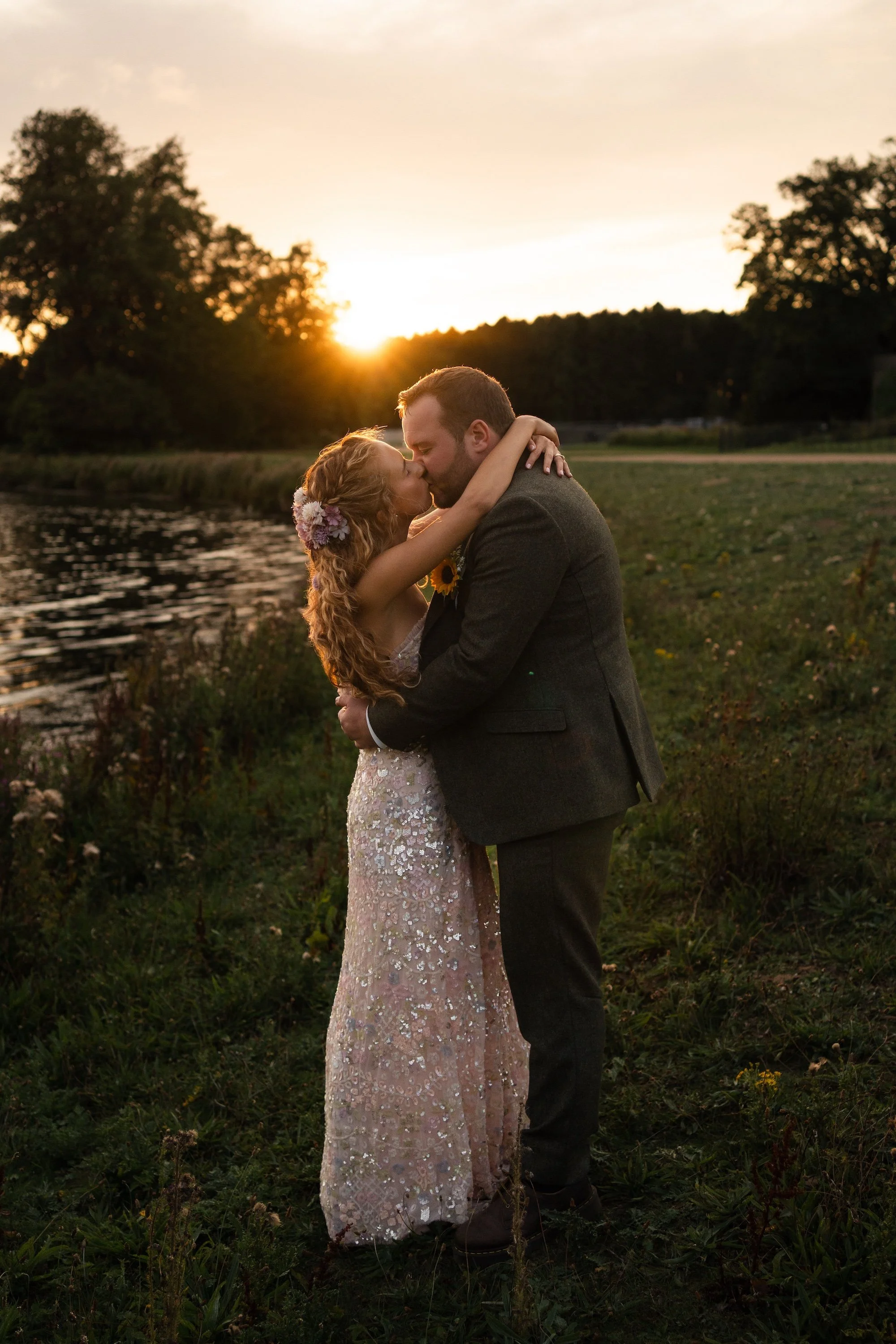 A couple sharing a kiss at sunset outdoors, the woman with flowers in her hair and the man in a suit, embracing by a river with trees in the background. Hardwick Hall Hotel. 