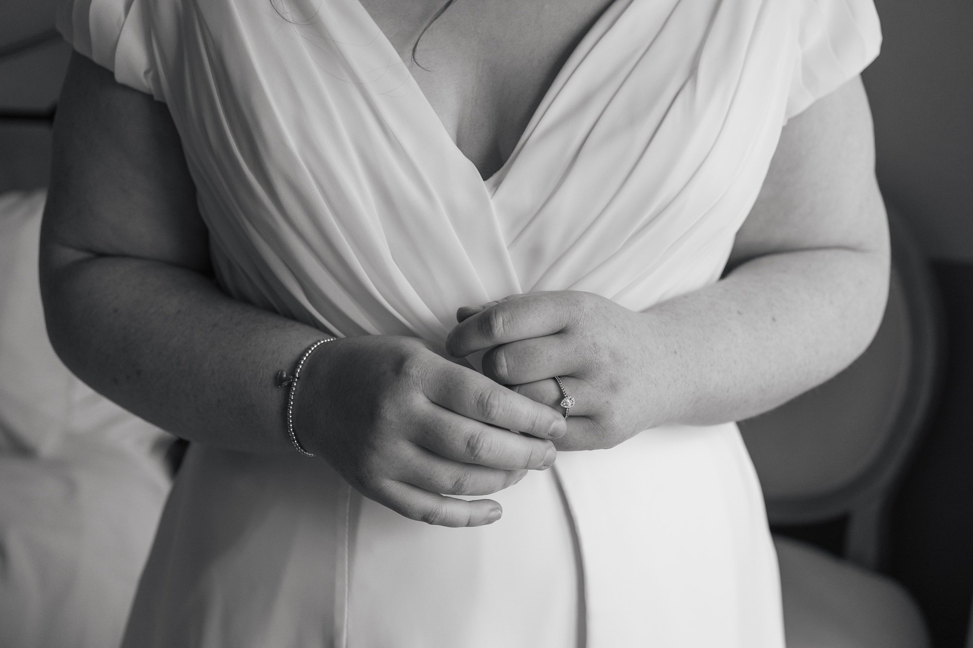 Close-up of a woman wearing a white dress, showing her hands adorned with a ring and a bracelet, resting on her stomach.