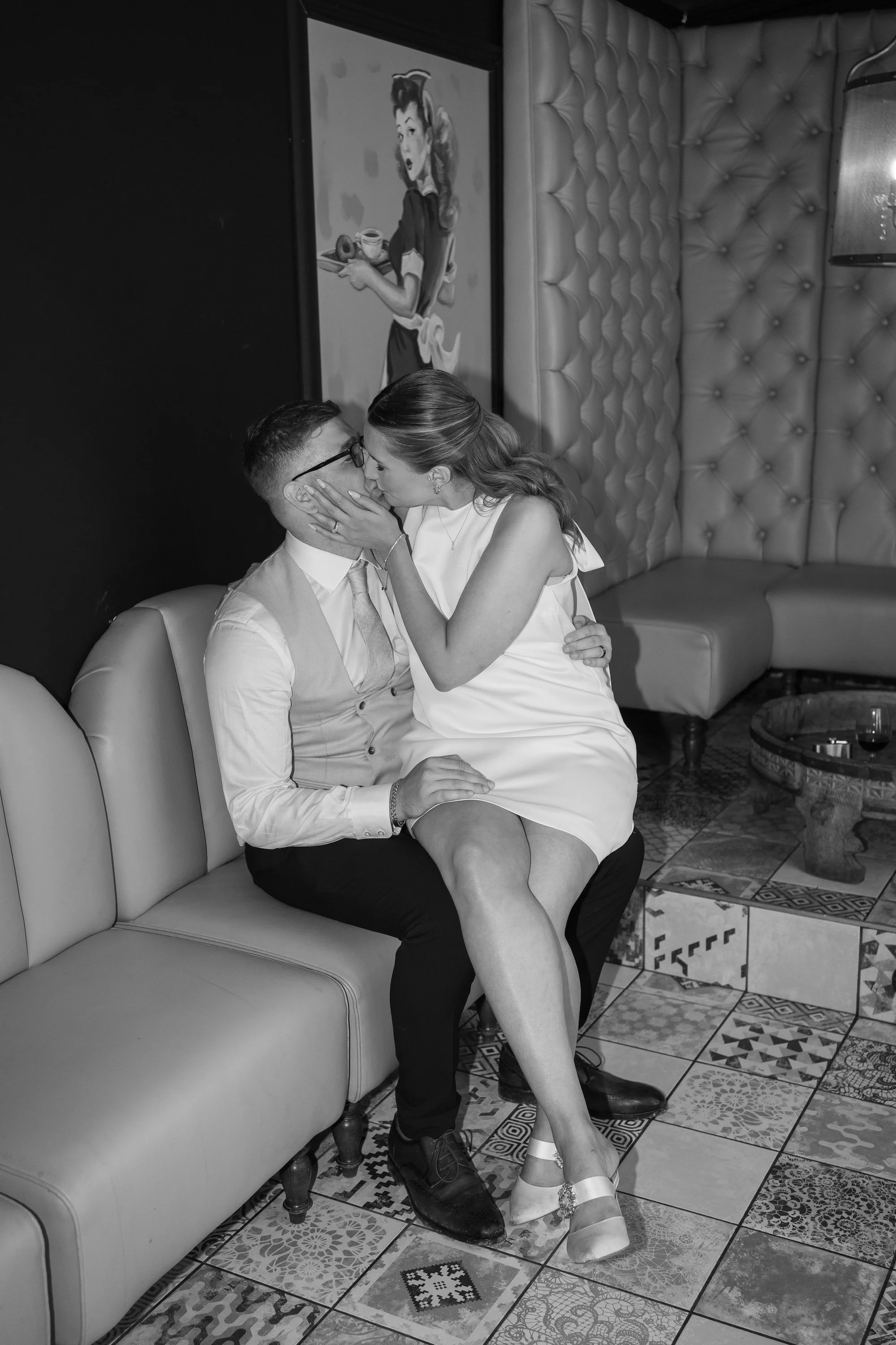 A black-and-white photo of a couple sitting on a sofa, sharing a kiss. The woman is wearing a sleeveless dress and high heels, while the man is dressed in a shirt, vest, and trousers. They are in a decorated interior with patterned tiled floors and a