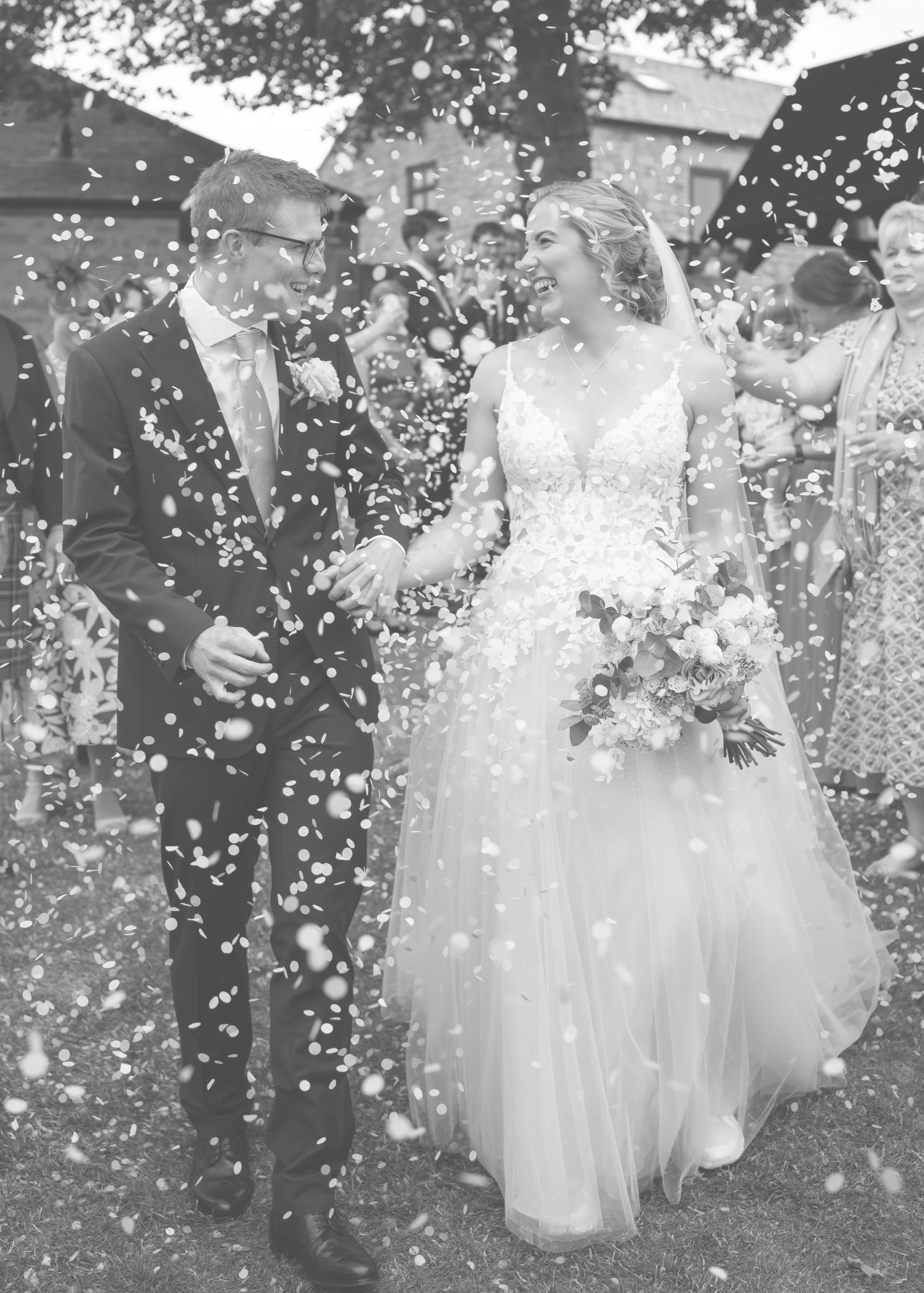 Black and white photo of a bride and groom walking outdoors, smiling, holding hands, celebrating their wedding with confetti, surrounded by guests. South Causey Inn wedding photography.