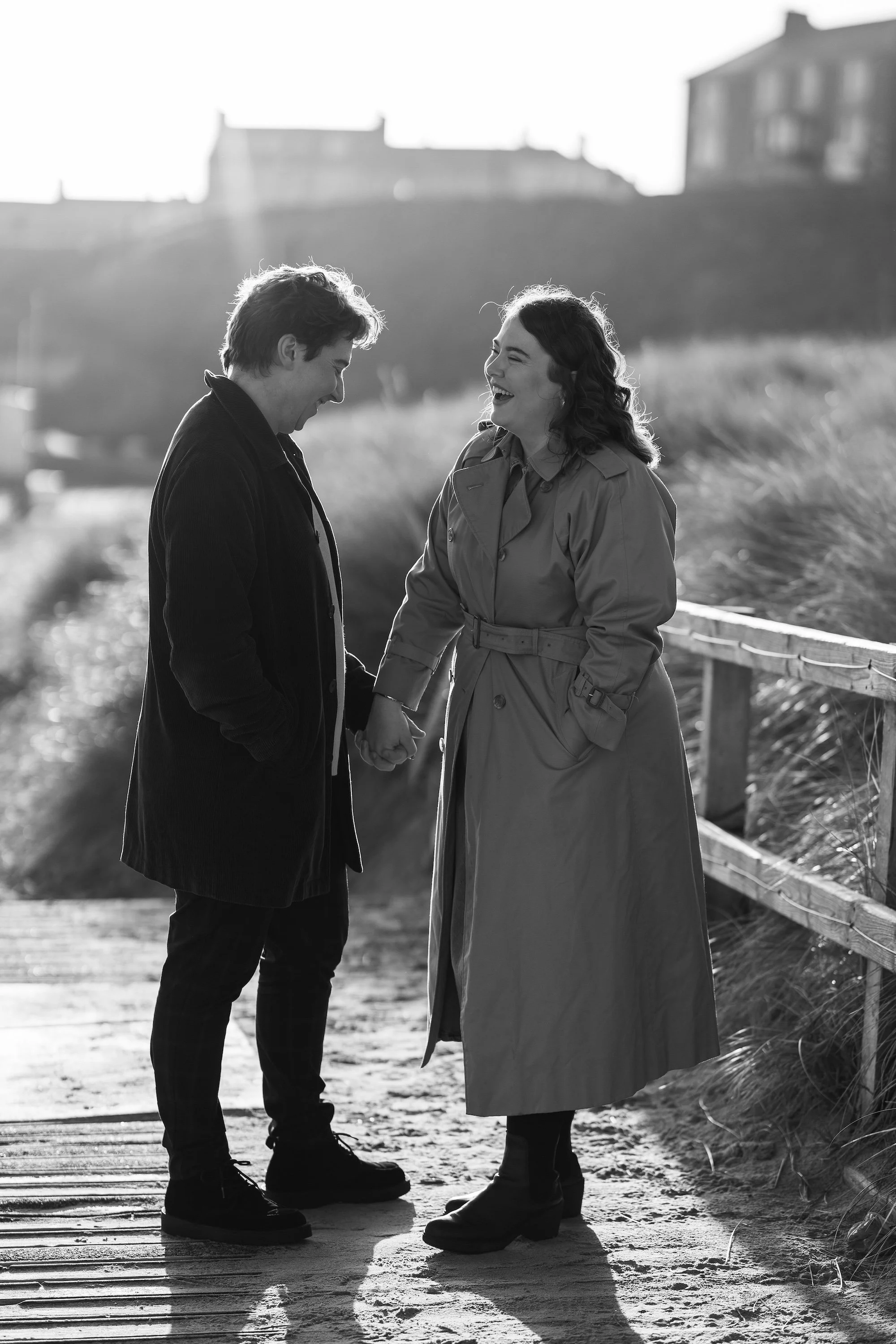 A black and white photo of a man and woman standing on a sandy path outdoors, holding hands, smiling, and sharing a happy moment. The man has dark hair and is dressed in dark clothing, while the woman has dark curly hair and is wearing a long trench 