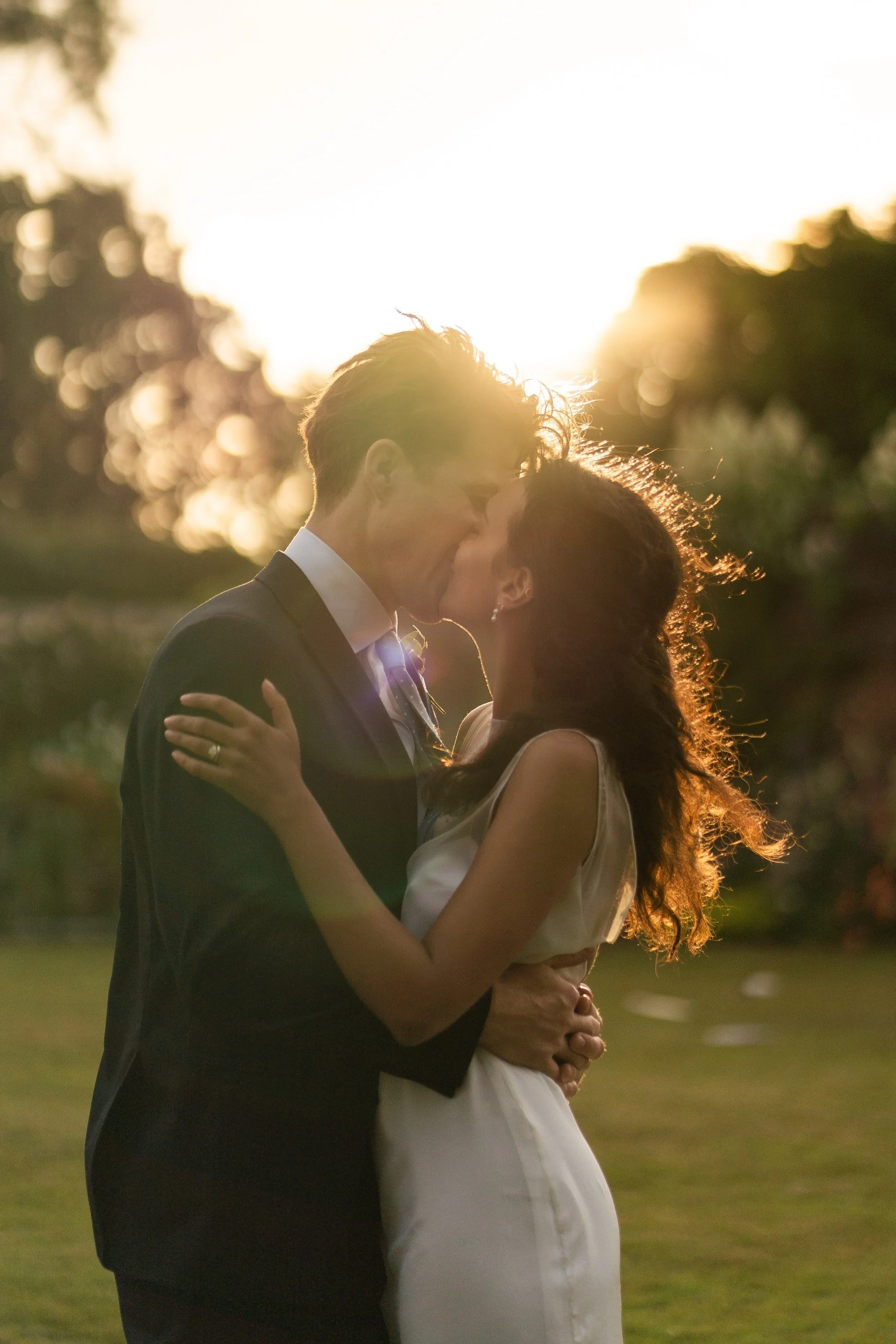 A couple in wedding attire kissing outdoors at sunset, with trees in the background. Whalton Manor Gardens.