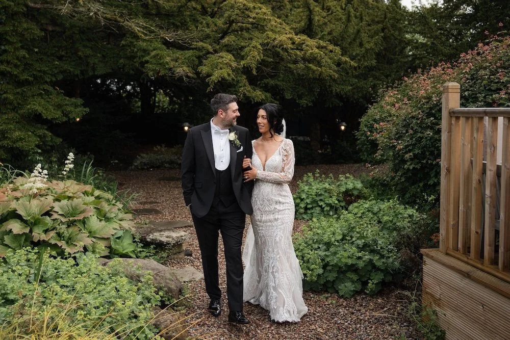 A bride and groom walking together outdoors on their wedding day, surrounded by green foliage and garden plants. Horton grange wedding photos. 