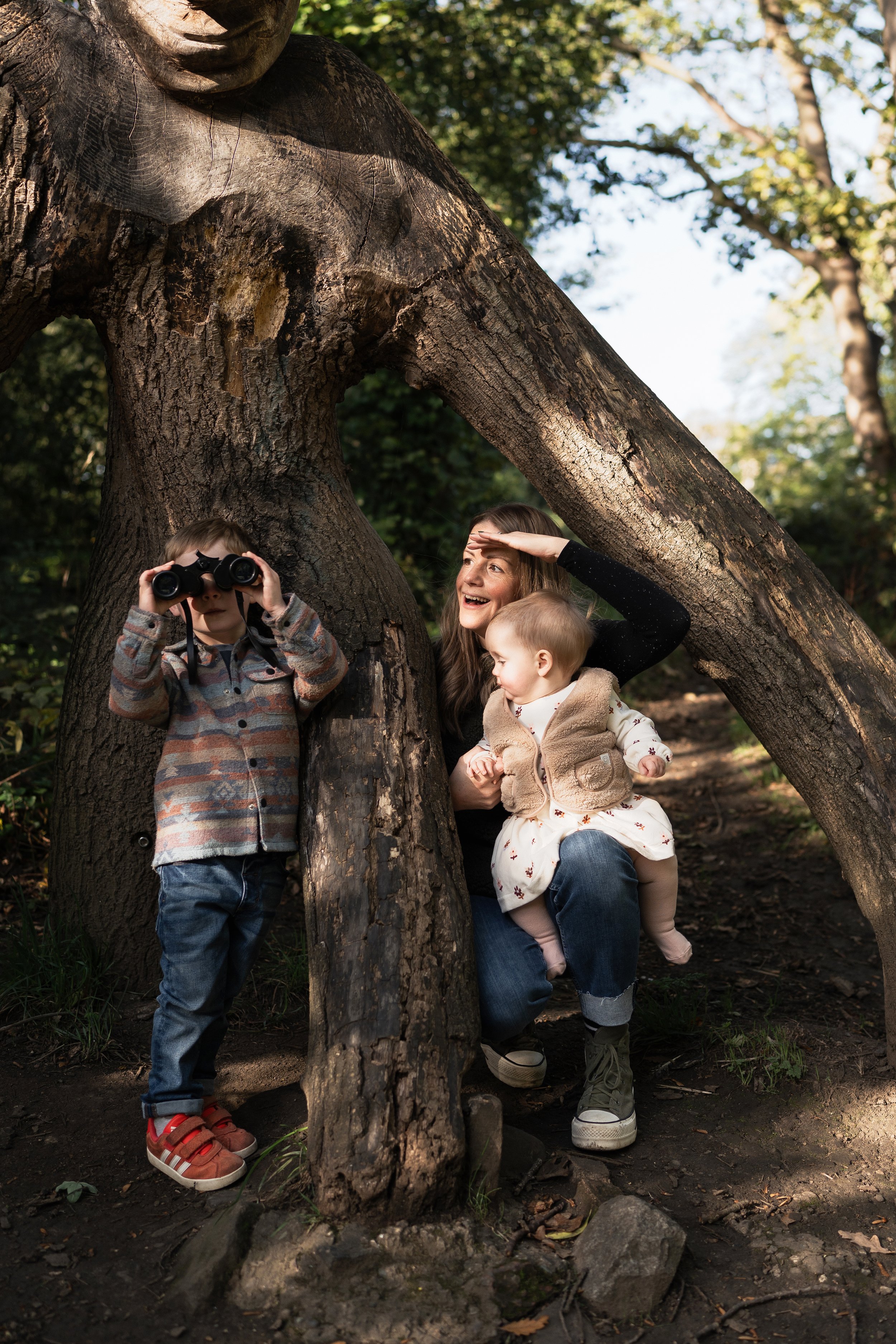 A woman with two children, a boy with binoculars and a girl, play behind a large fallen tree in a forest.