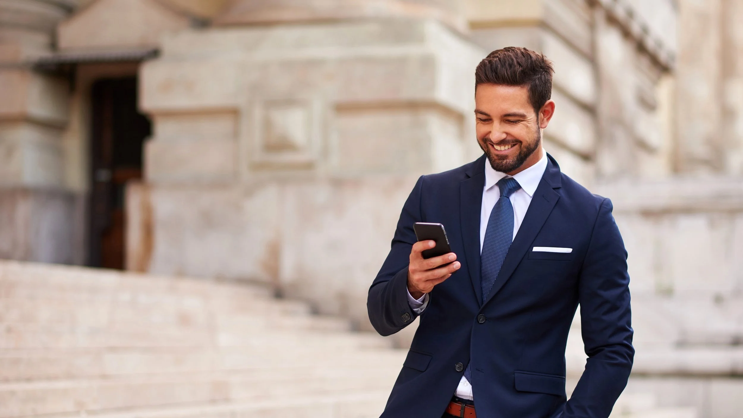 A man in a navy blue suit with a white shirt and a blue tie is smiling while looking at his smartphone, standing outside a historic building with geometric, stone architecture.