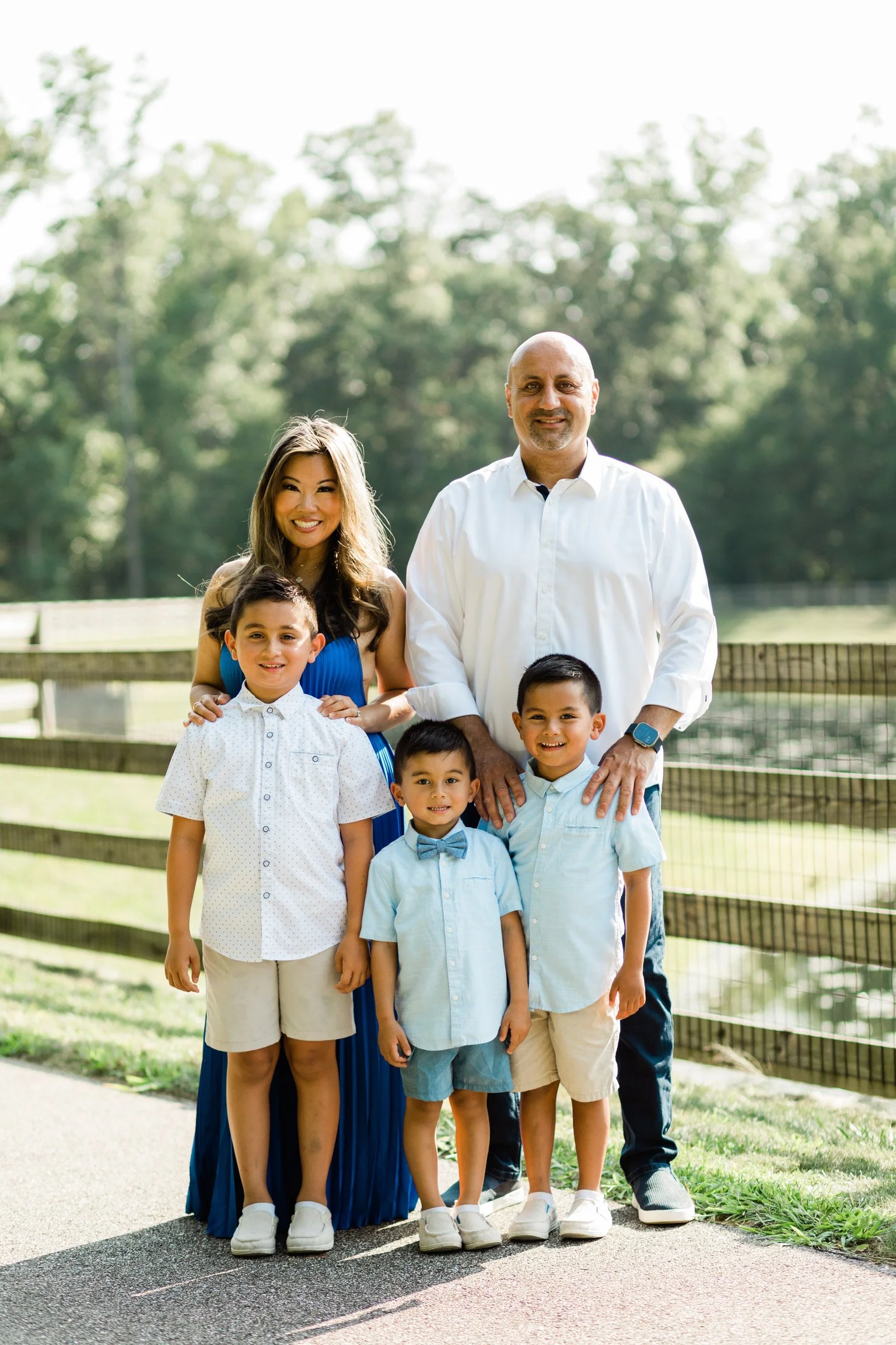 Lisa Chiya, wearing a blue dress, Page Tofighi, wearing a white dress shirt, and their three children born via IVF and surrogacy, stand outdoors in front of a wooden fence and green trees on a sunny day, smiling at the camera.