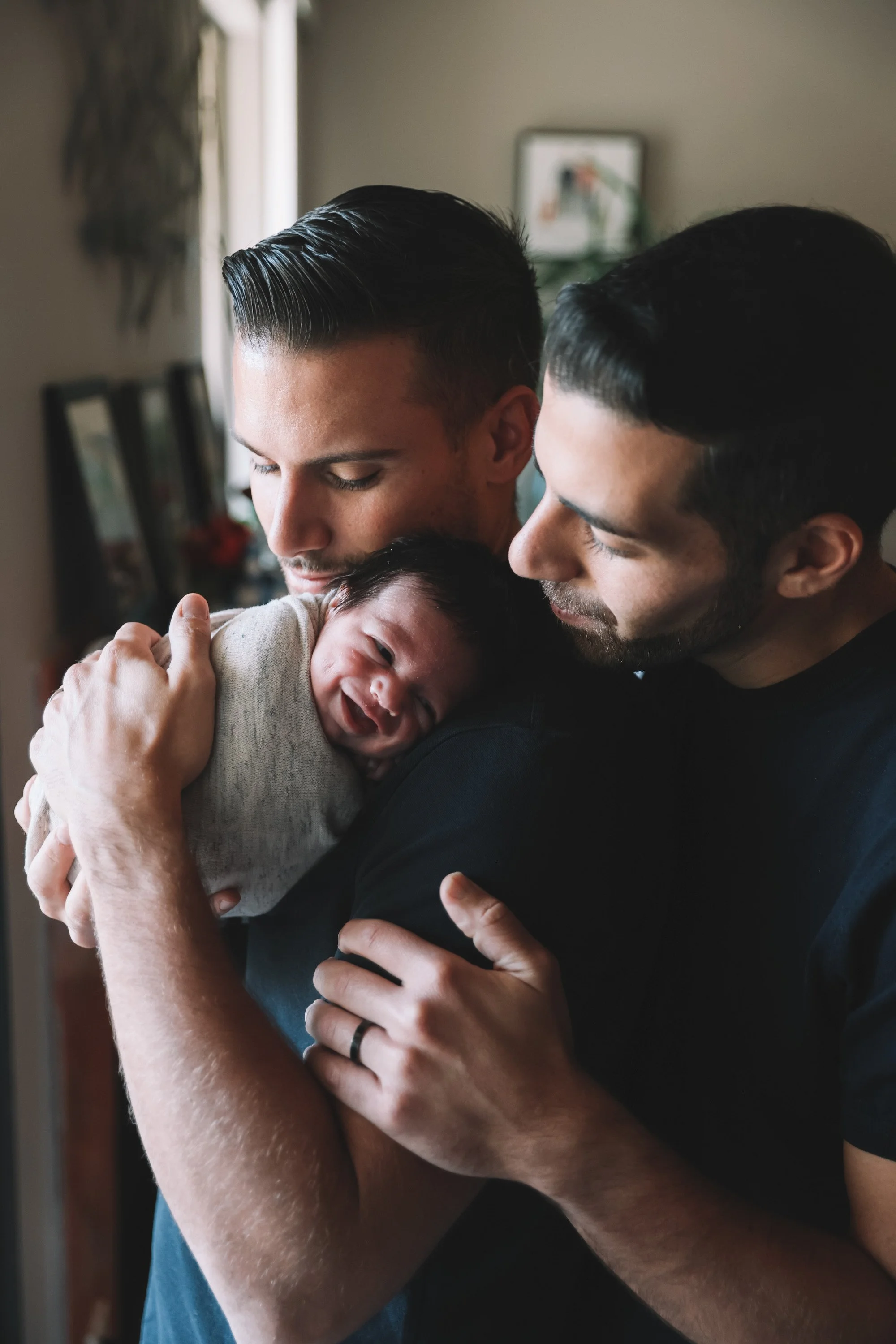 Two dark-haired men, Genesis Group intended parents, hold their smiling, swaddled baby born via IVF and surrogacy together in their home, with a blurred background.