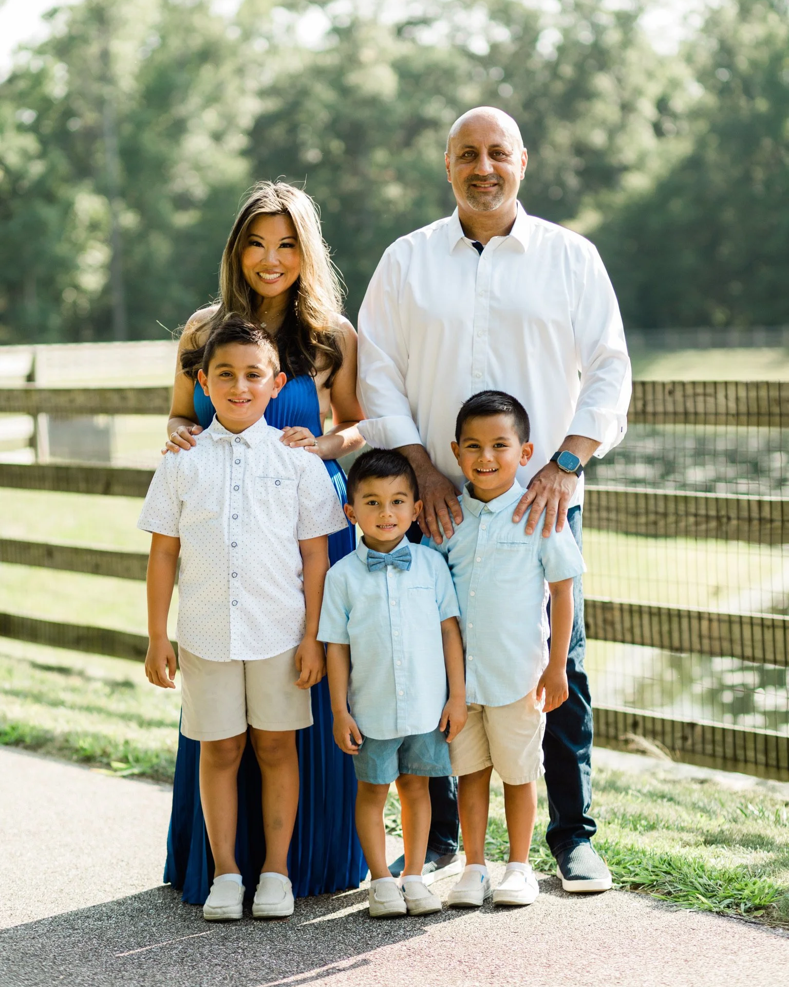 A family of five stands outdoors in front of a wooden fence, with trees in the background. The family includes a woman, a bald man, and four young boys. They are Lisa Chiya, Page Tofighi, and kids, all smiling and dressed in light, casual clothing.