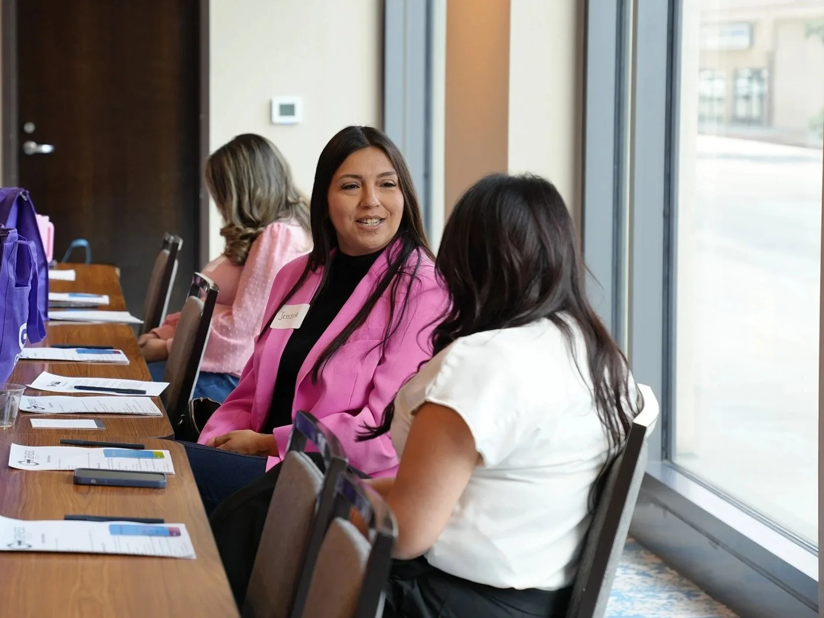 Two women, The Genesis Group surrogate Jessica and senior case manager Heidi Morales, are sitting at a conference table engaging in conversation. There are pamphlet, notebooks, and gift bags on the table, and a window with a view of the city outside.
