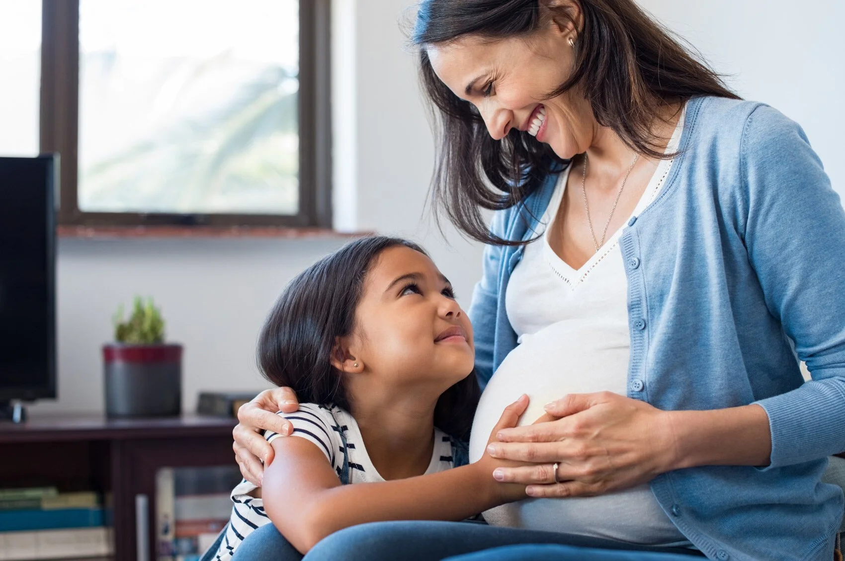 A pregnant woman smiling and holding her belly, being hugged by a young girl looking up at her in a cozy room with a window, a plant, and a computer in the background.