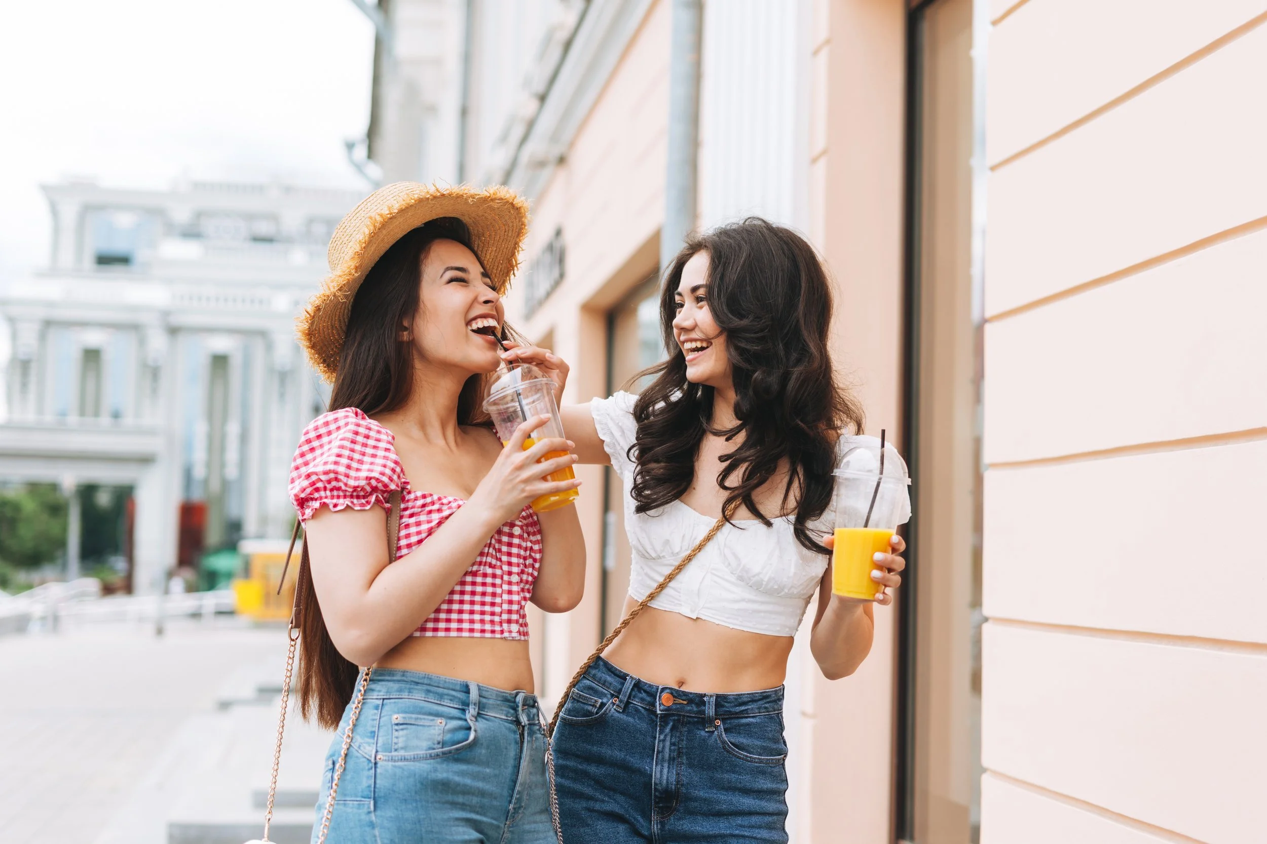 Two women enjoying drinks and laughing outdoors on a city street, one wearing a straw hat and red checkered top, the other with long dark hair and a white crop top.