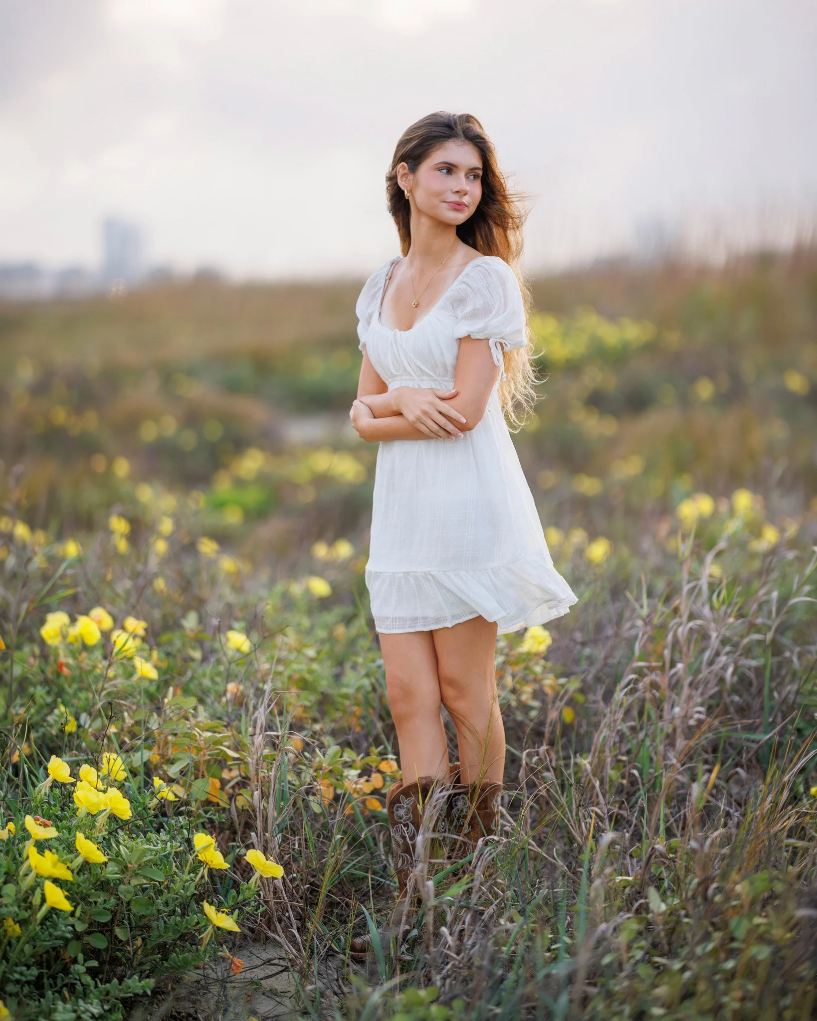 A young woman with long brown hair standing in a field of yellow flowers and green foliage, wearing a white dress, with a cityscape in the background under an overcast sky.