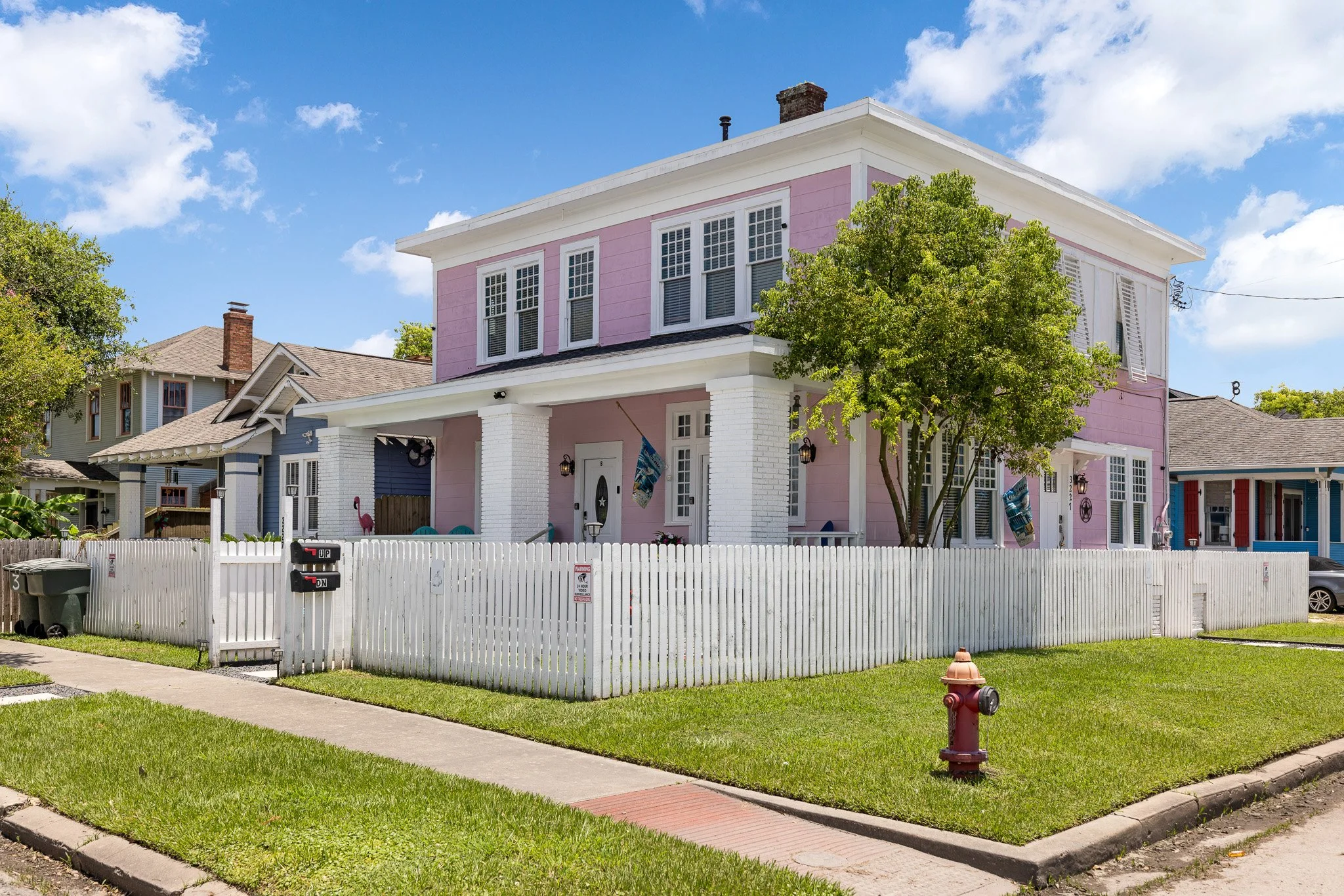 Colorful two-story house painted pink and white with a front porch, surrounded by a white picket fence, green lawn, and a bright blue sky with clouds.