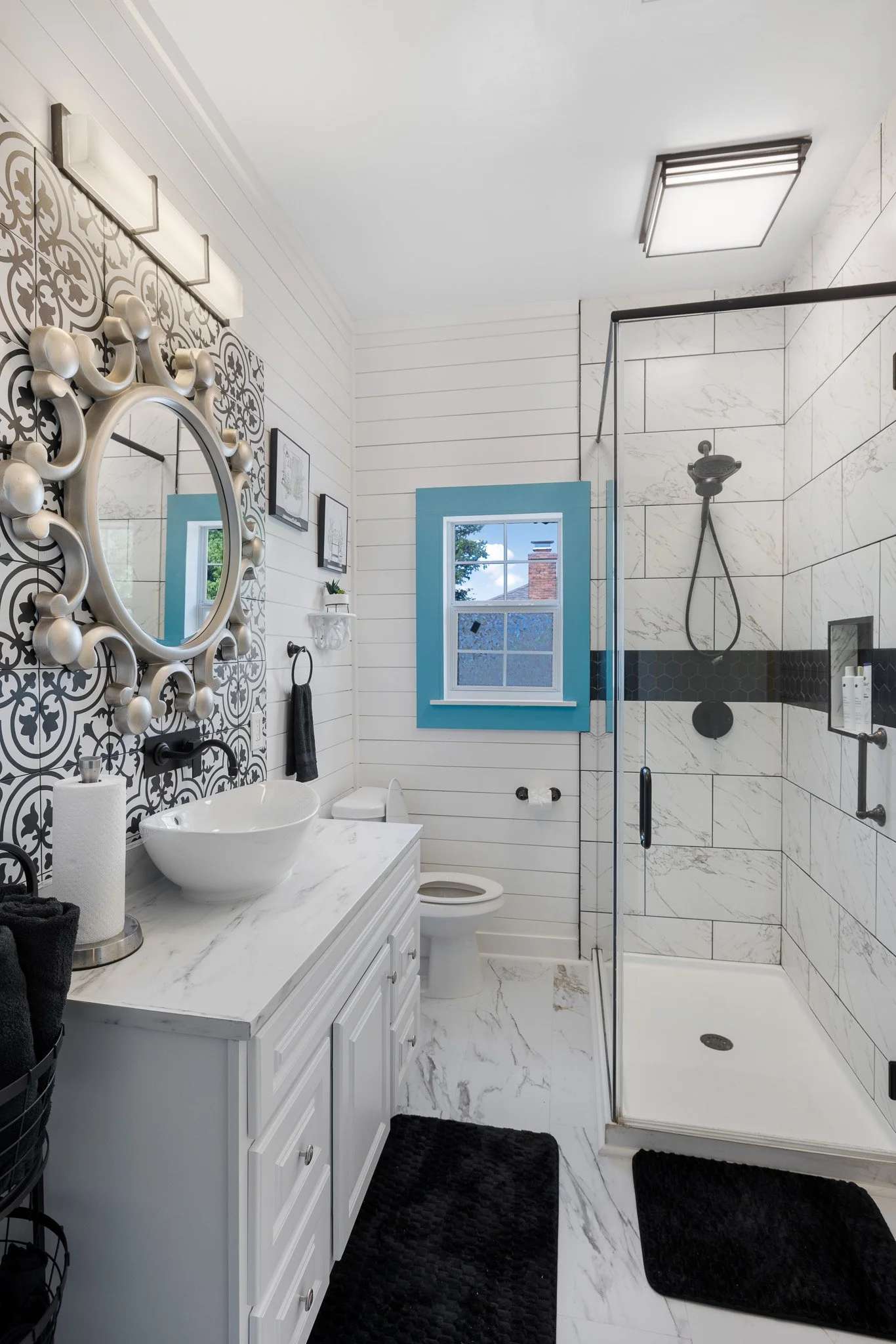 Modern white bathroom with a patterned mirror, white vanity with a vessel sink, black towels, a walk-in shower with white marble tiles and a black horizontal accent, a window with blue trim, and black bathroom mats.