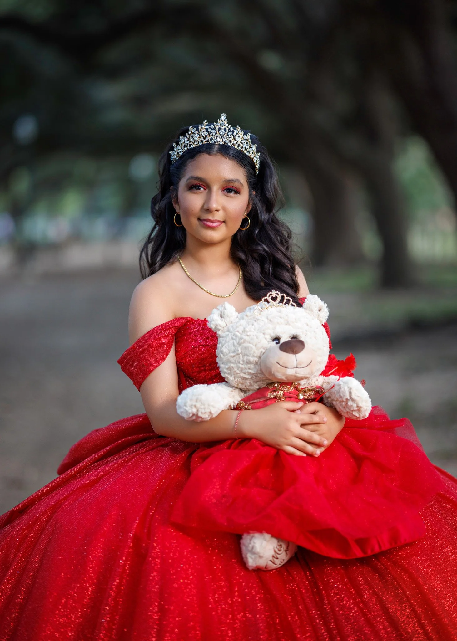 Young woman wearing a red gown and tiara, holding a teddy bear, outdoors with trees in the background.