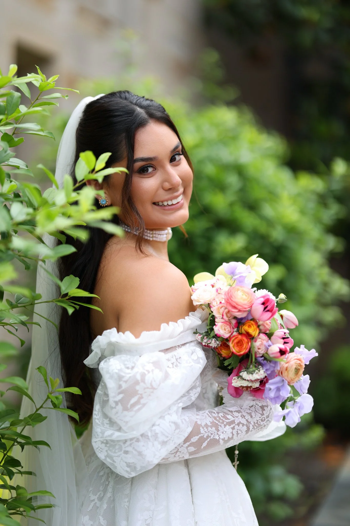 A bride with long dark hair wearing a white off-shoulder lace dress, holding a colorful bouquet of pink, orange, and purple flowers, smiling with greenery in the background.