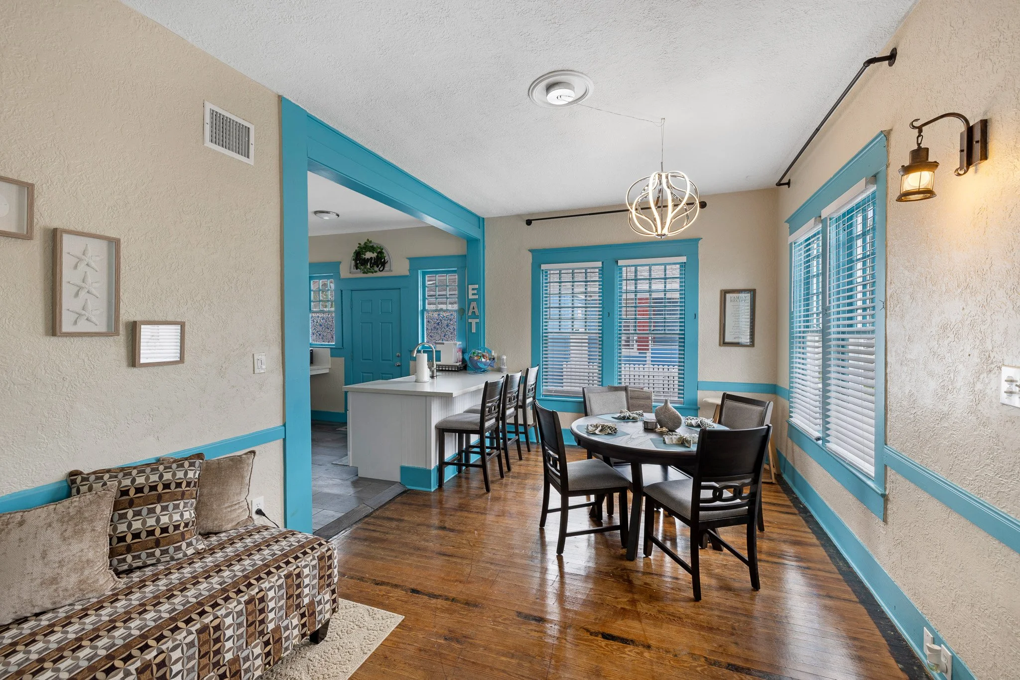 A dining room with beige textured walls, blue trim around the windows and doorway, a round dark wood dining table with six chairs, and a sofa with patterned cushions. There are three windows with blinds, a modern chandelier hanging from the ceiling,