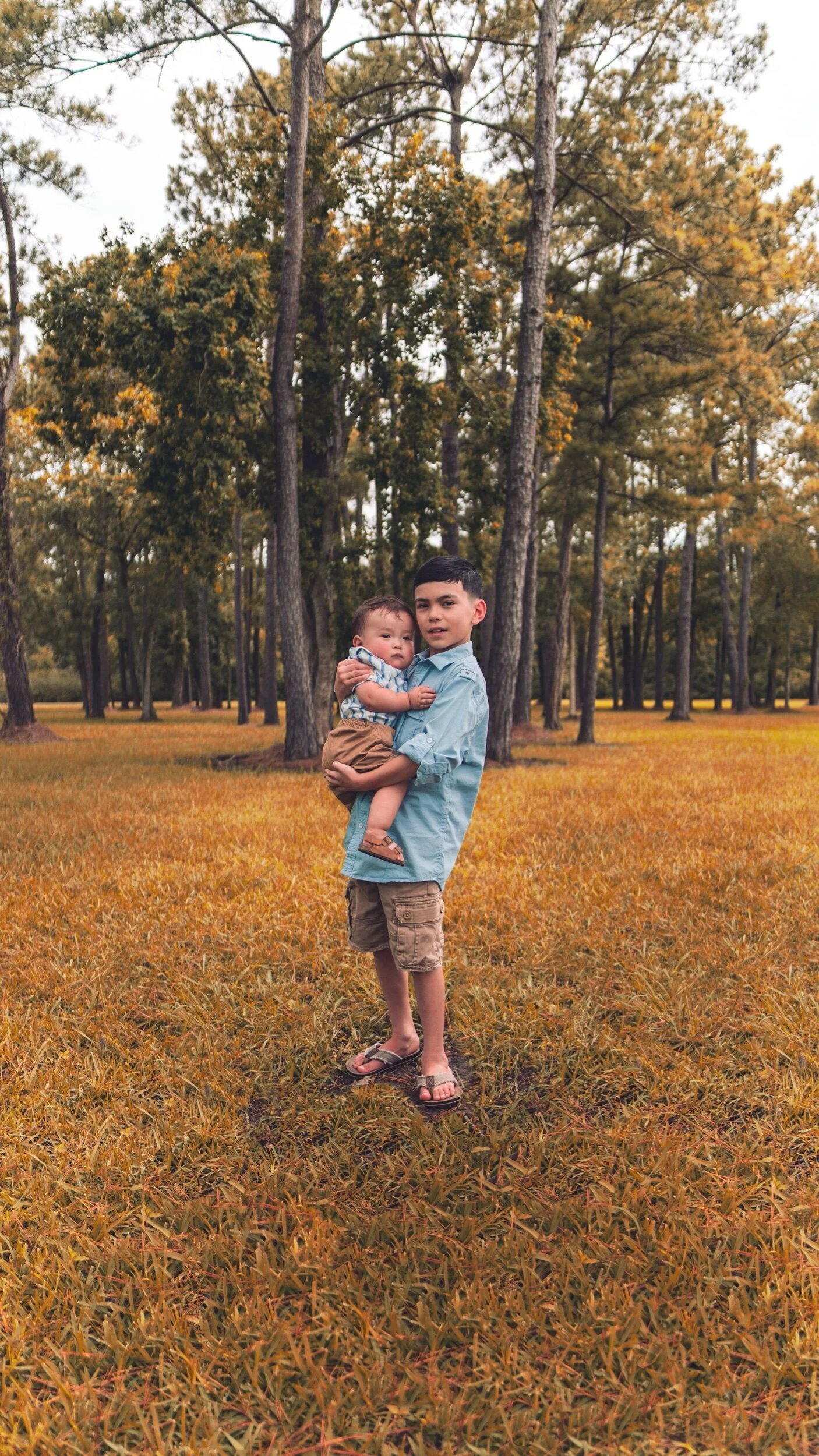 A young boy with short dark hair holding a toddler in a park with tall trees and yellow-orange fall foliage.