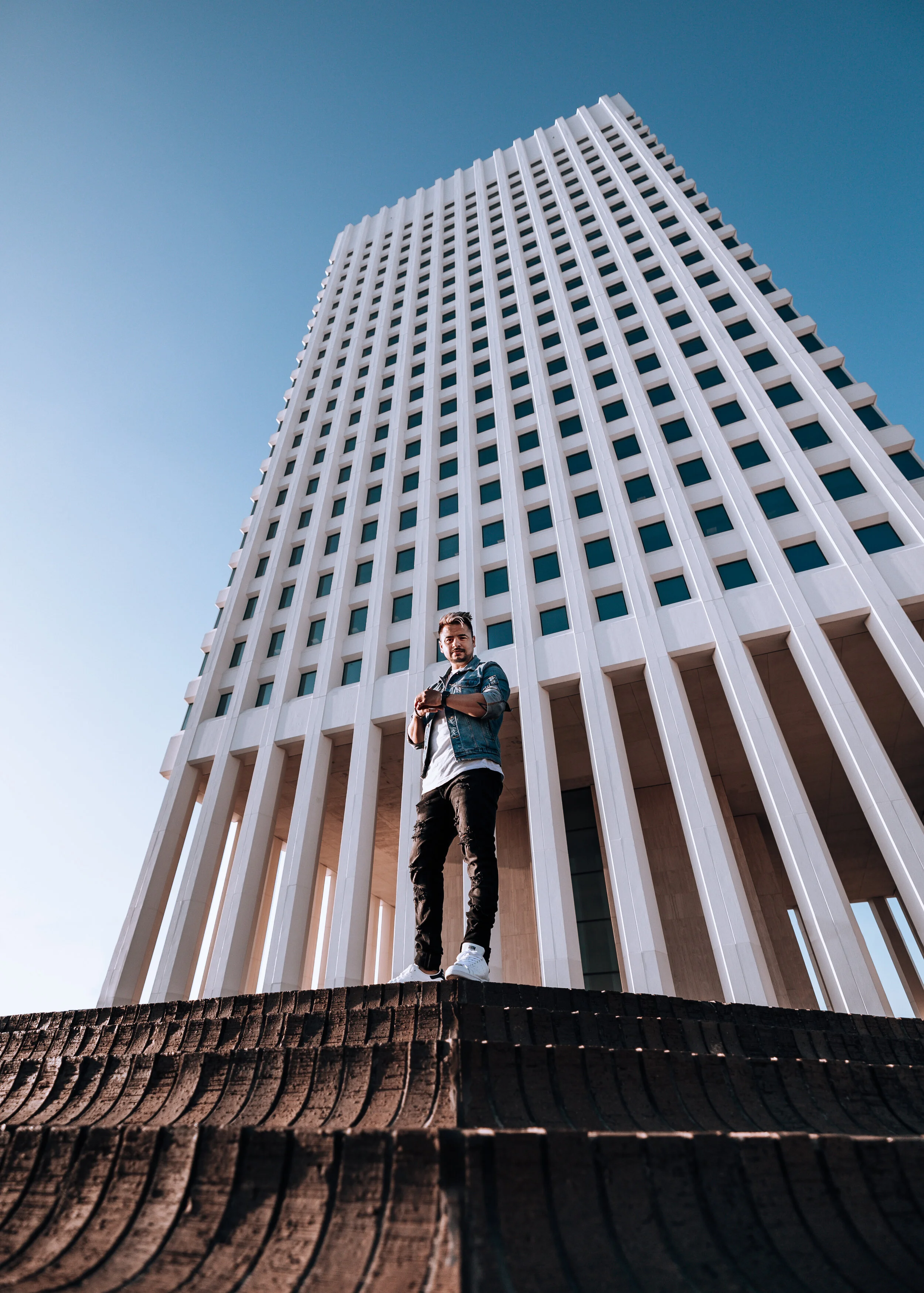A man standing outdoors in front of a modern tall building with white vertical stripes, under a clear blue sky.