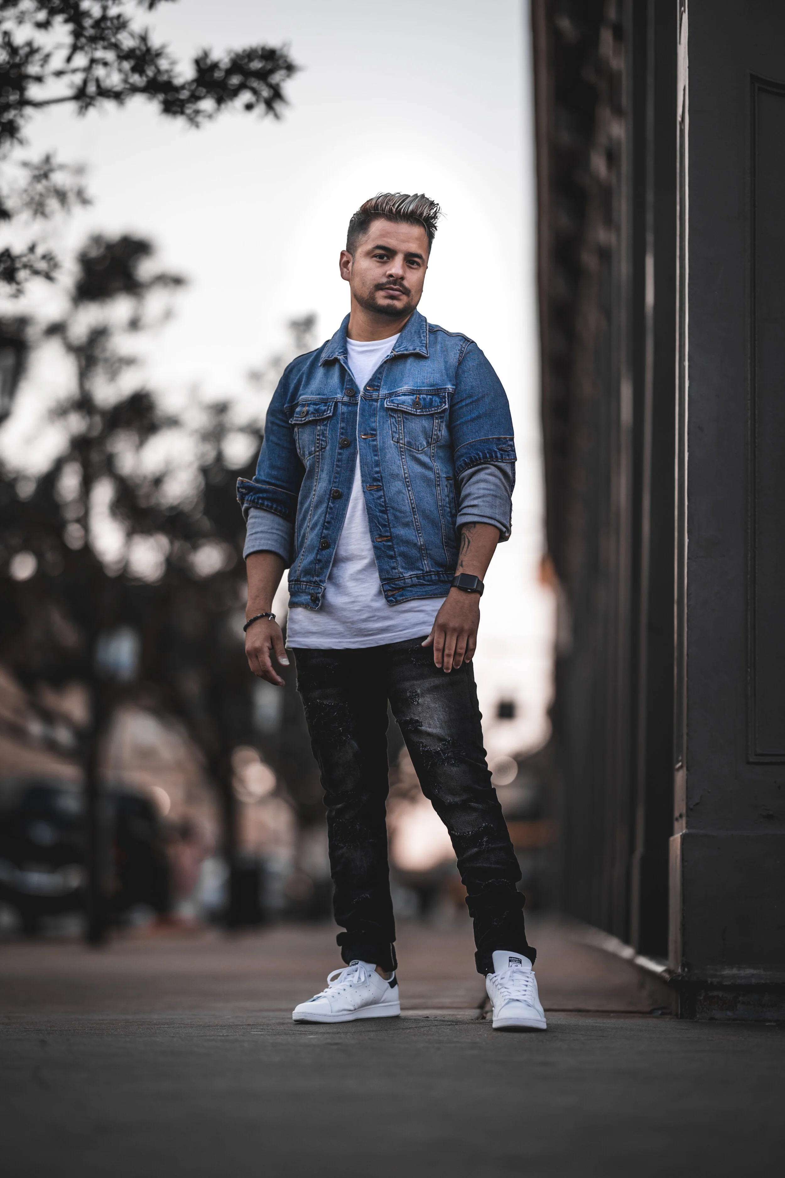 A young man in a denim jacket, black jeans, and white sneakers standing on a city street near a building during dusk.