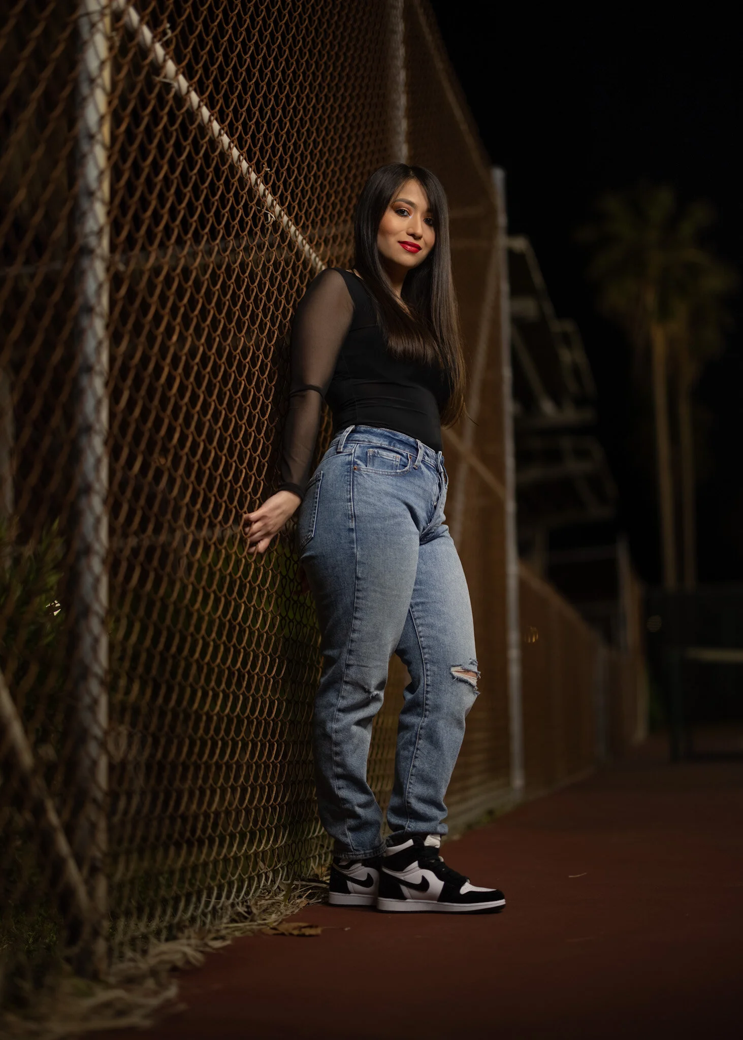 Young woman with long dark hair wearing a black top with sheer sleeves, blue ripped jeans, and black and white athletic shoes, standing by a chain-link fence at night.