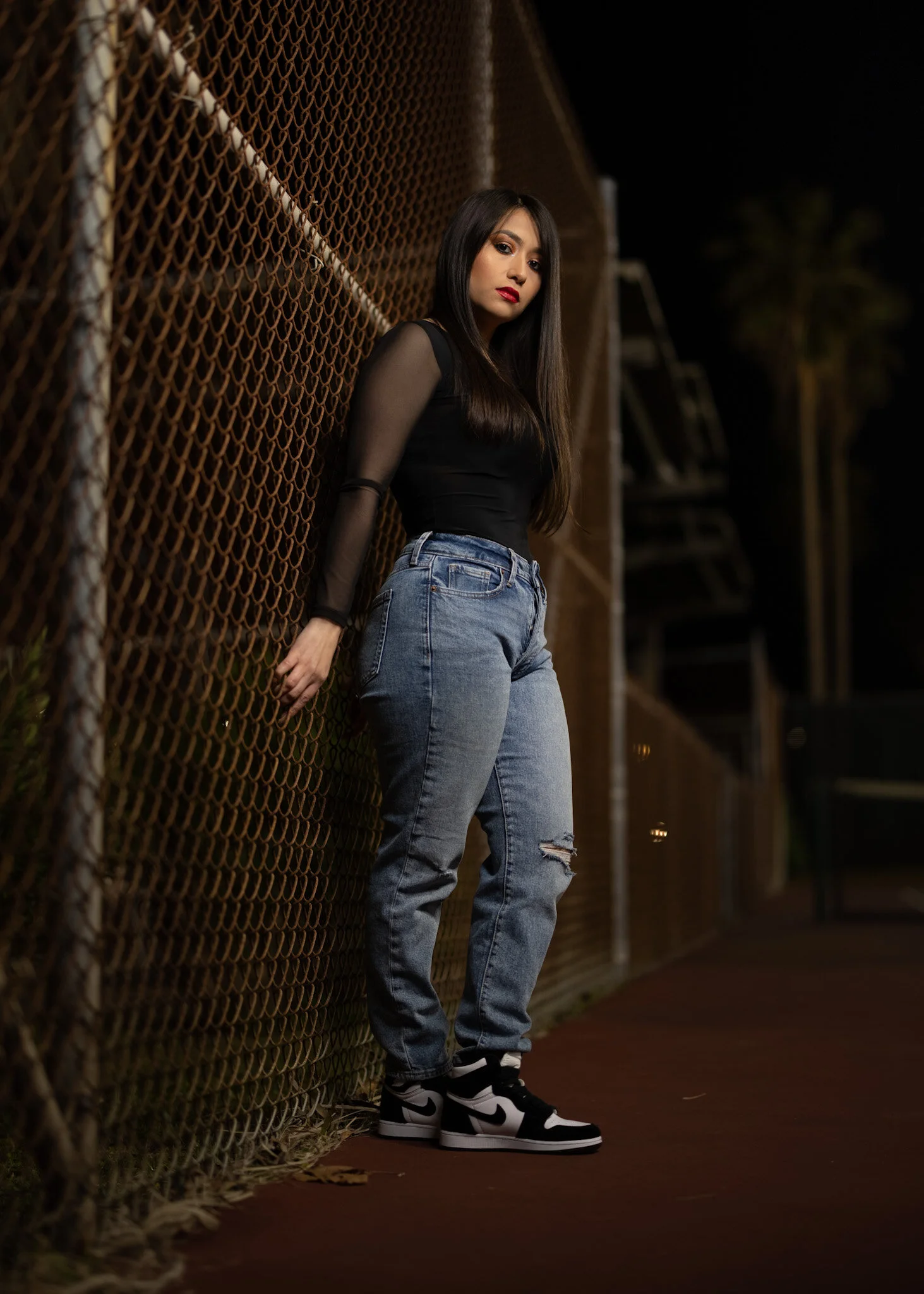 A young woman with long dark hair and red lipstick stands against a chain-link fence at night, wearing a black top with sheer sleeves, blue ripped jeans, and black and white sneakers.