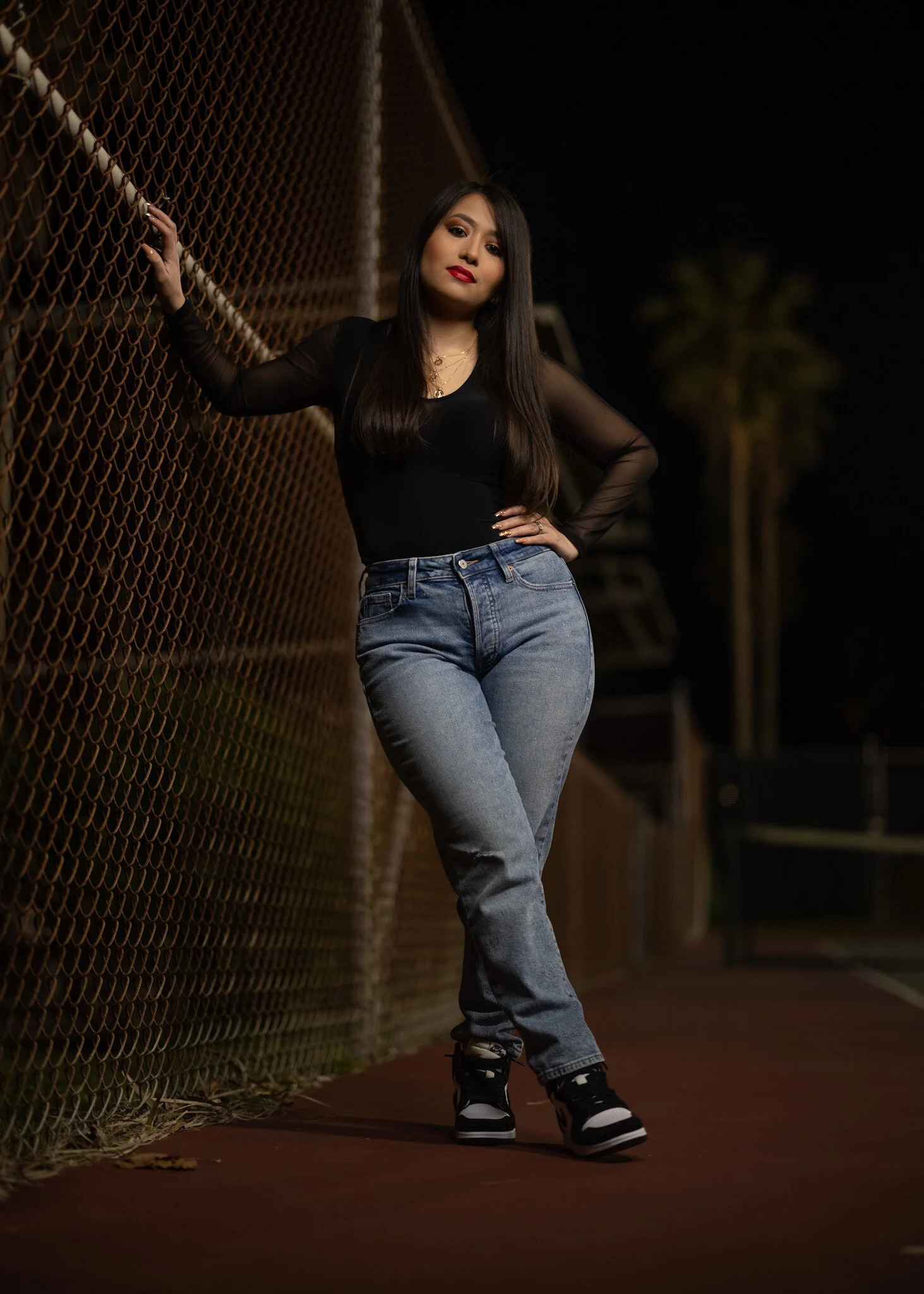 A young woman with long dark hair and red lipstick wearing a black sheer long sleeve top and high-waisted blue jeans standing against a chain-link fence at night.