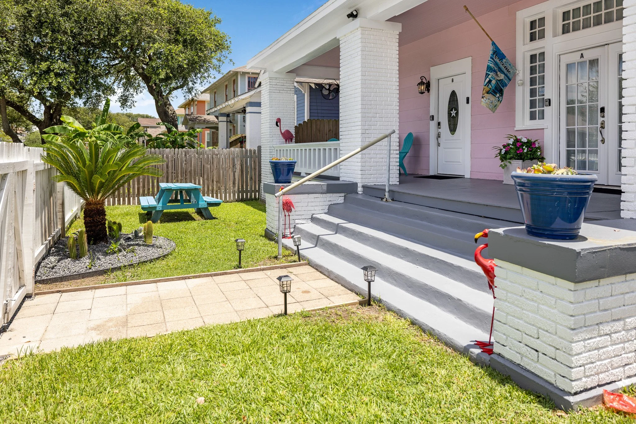 Front porch and yard of a house with pink and white exterior, decorated with pink flamingo lawn ornaments, potted plants, a blue picnic table, and tropical plants in a garden bed.