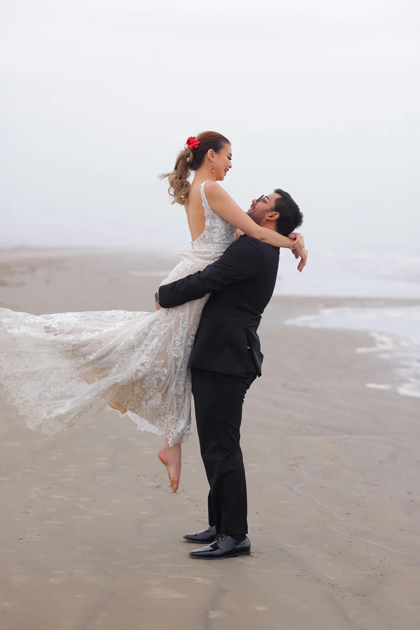 A couple on the beach, the man in a black suit lifting the woman in a wedding dress, she is smiling and looking at him, overcast sky with ocean in the background.