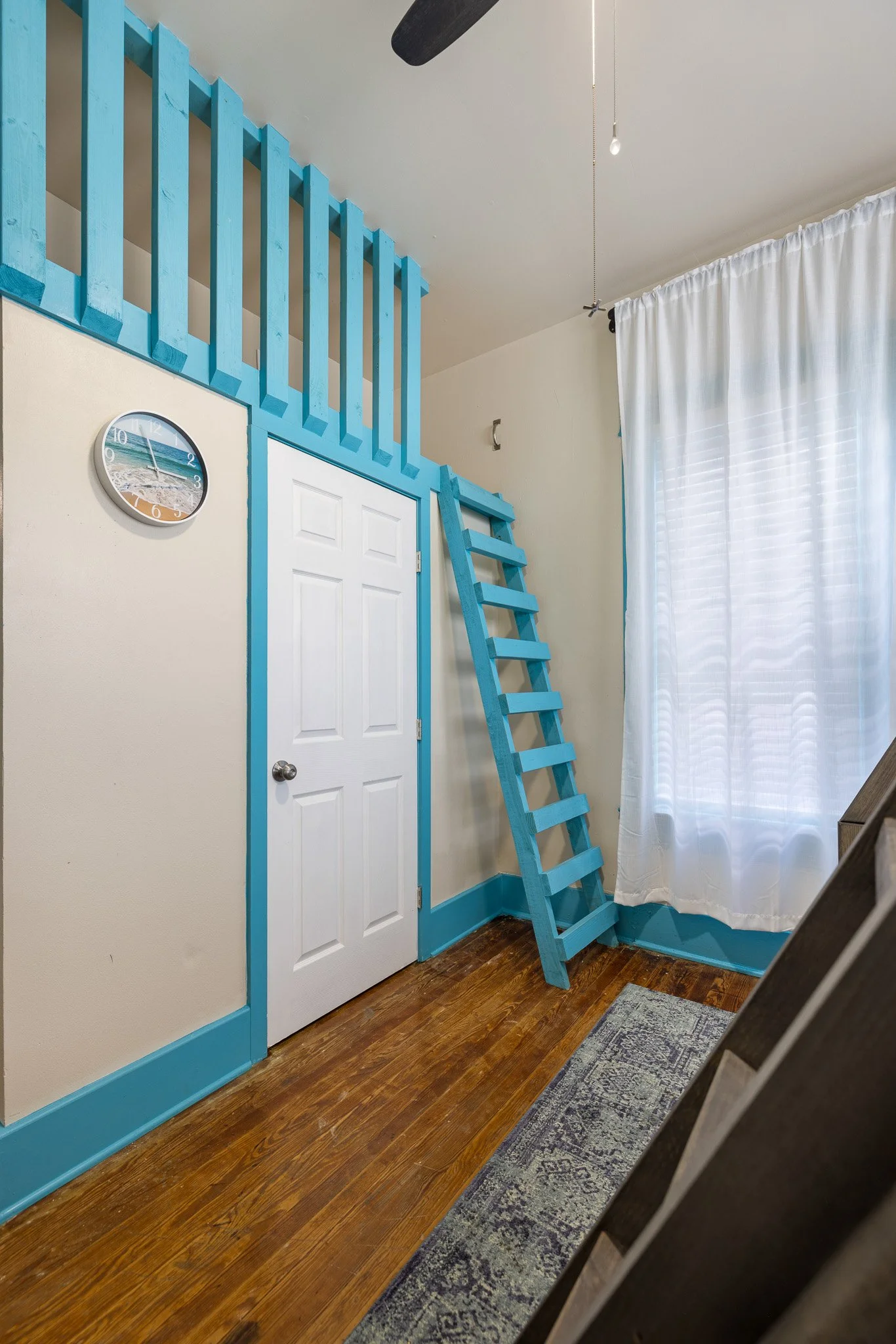 Room with a white door, light beige walls with blue trim, blue wooden ladder leading to a loft with a blue railing, a round wall clock showing 2:53, hardwood floor, white curtains over a window, and a small area rug.