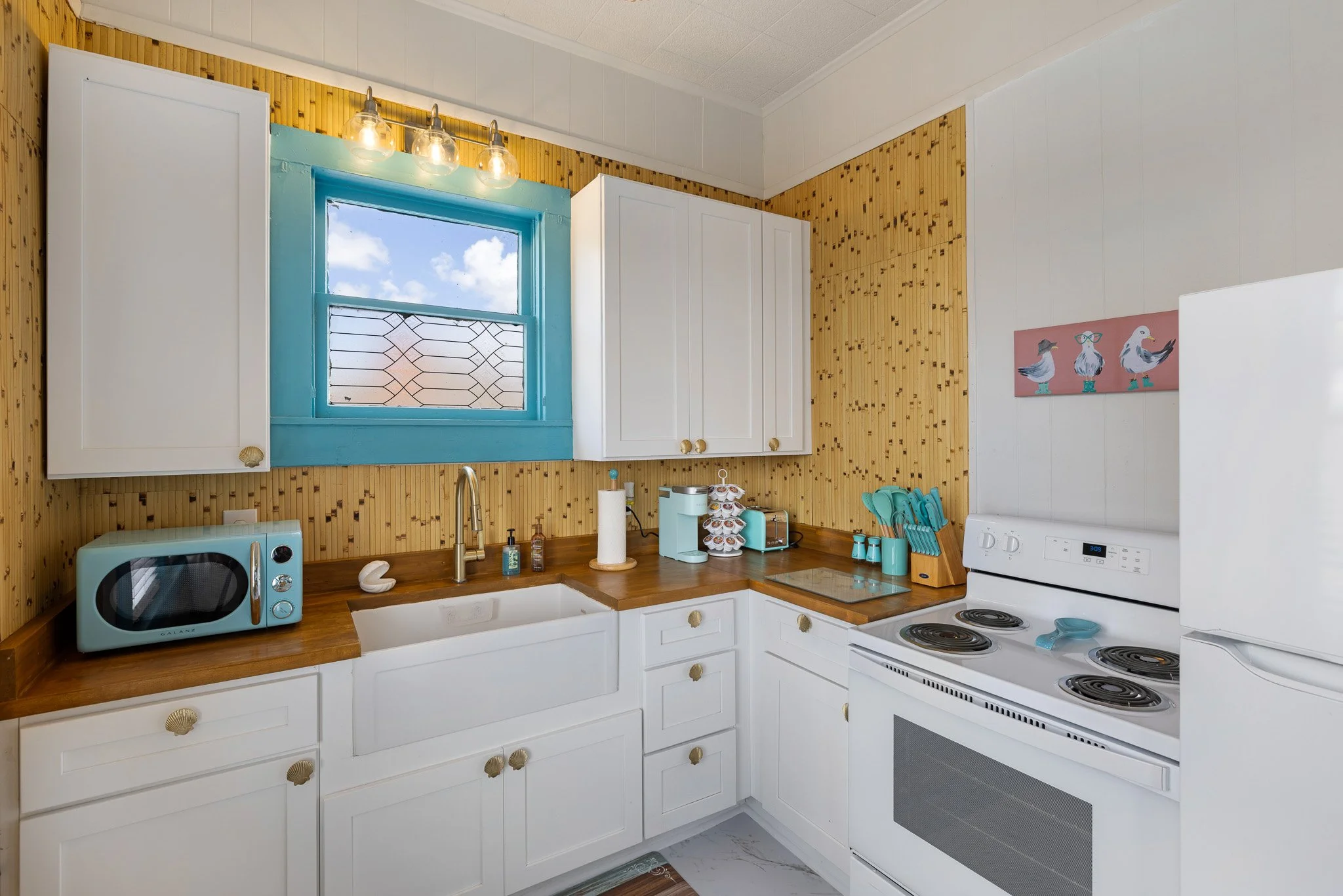 Kitchen with white cabinets, a wooden countertop, a window with blue frame, small appliances like microwave, coffee maker, and toaster, and a wall decorated with pink and white artwork of birds.