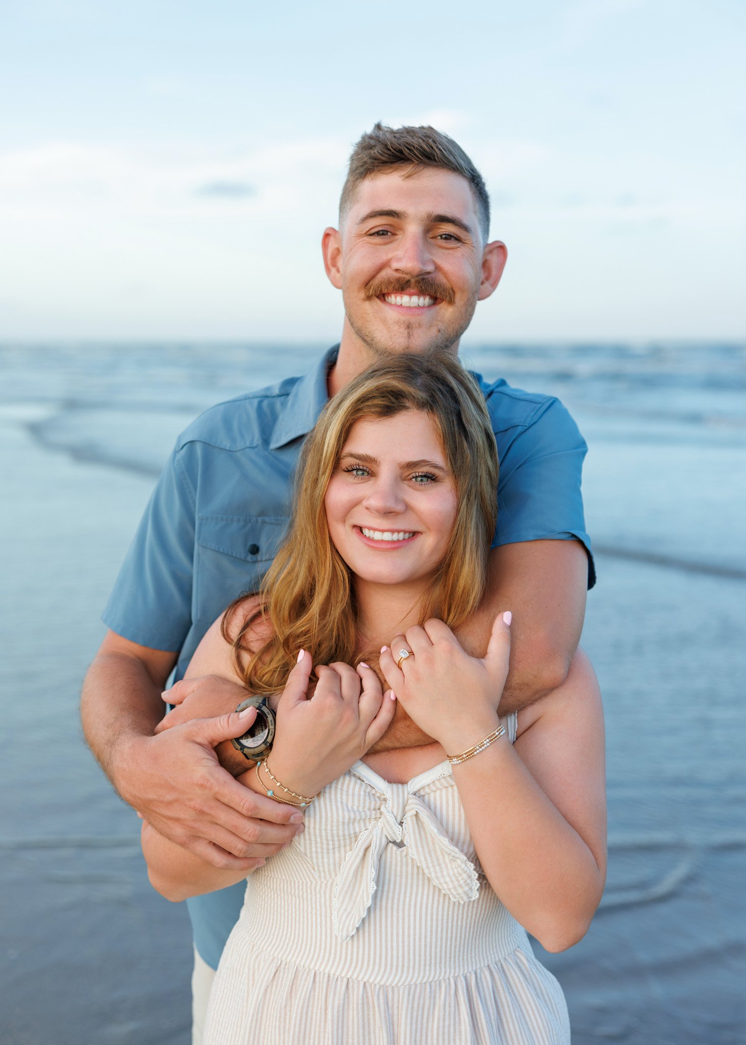 A smiling couple hugging on the beach with the ocean in the background.