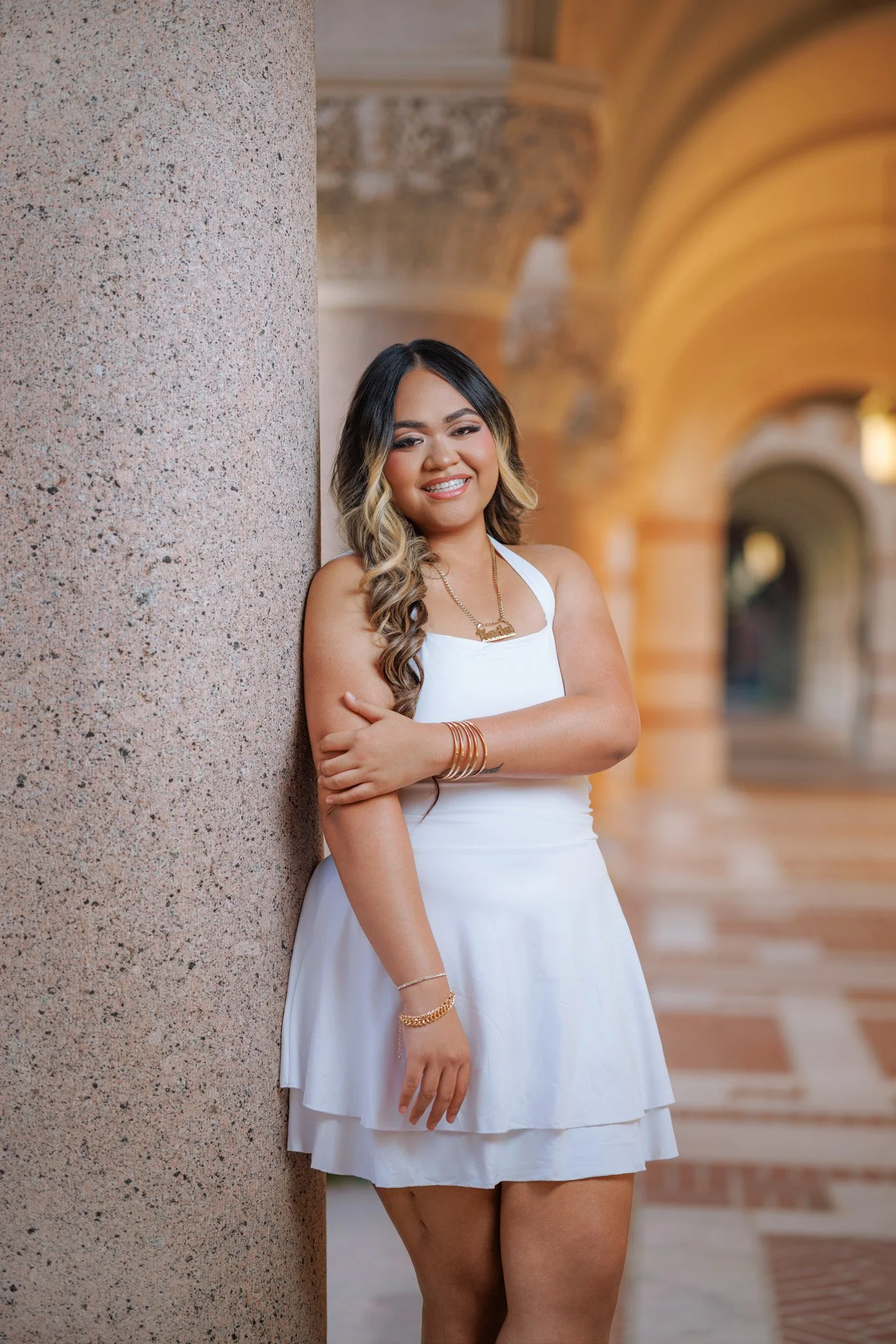A young woman with long, wavy hair standing next to a stone column in an architectural setting with arches, wearing a white sleeveless dress, gold jewelry, and smiling.