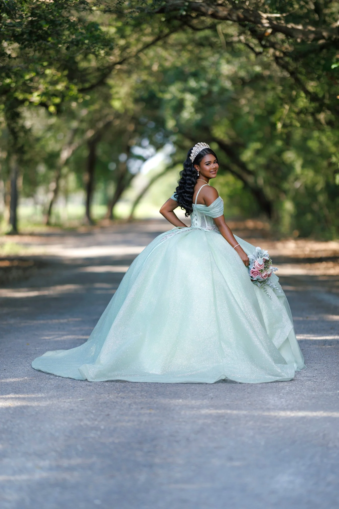 A young woman dressed as a princess in a light green ball gown poses outdoors under a canopy of trees, holding a bouquet of pink roses and smiling at the camera.