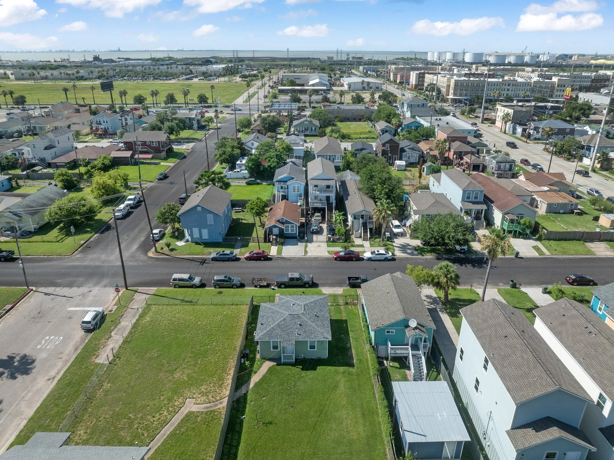 Aerial view of a residential neighborhood with houses, streets, and cars, with an industrial area and water in the background.