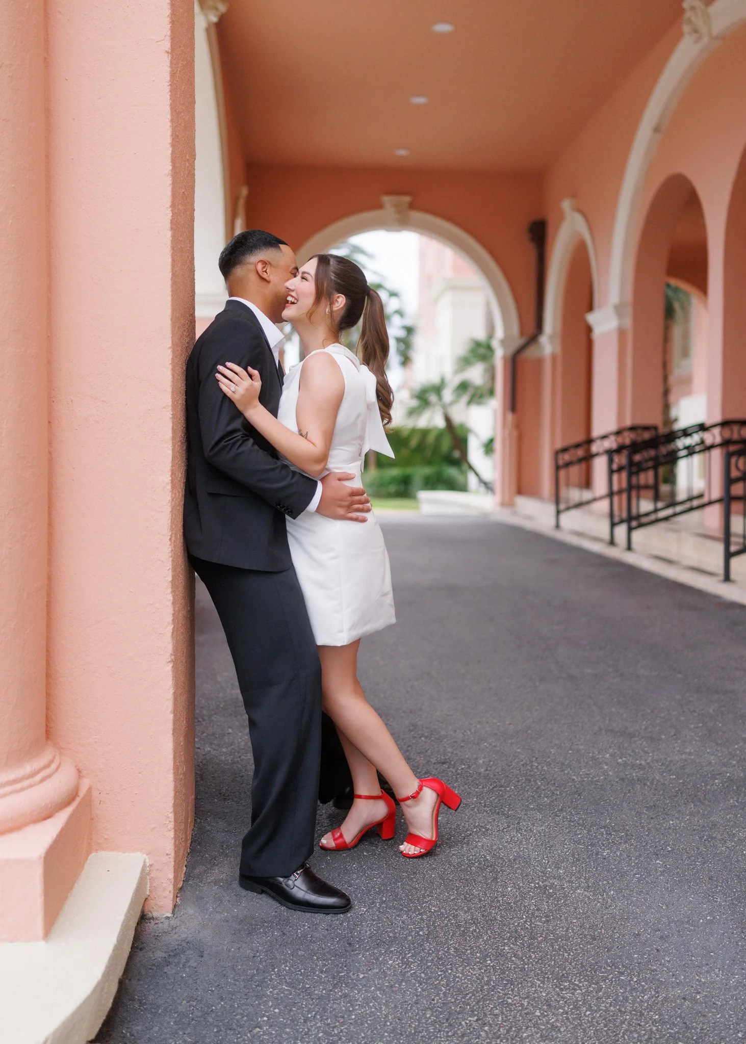 A couple dressed in formal attire standing in an archway, embracing and about to kiss, with the woman smiling and wearing red heels.
