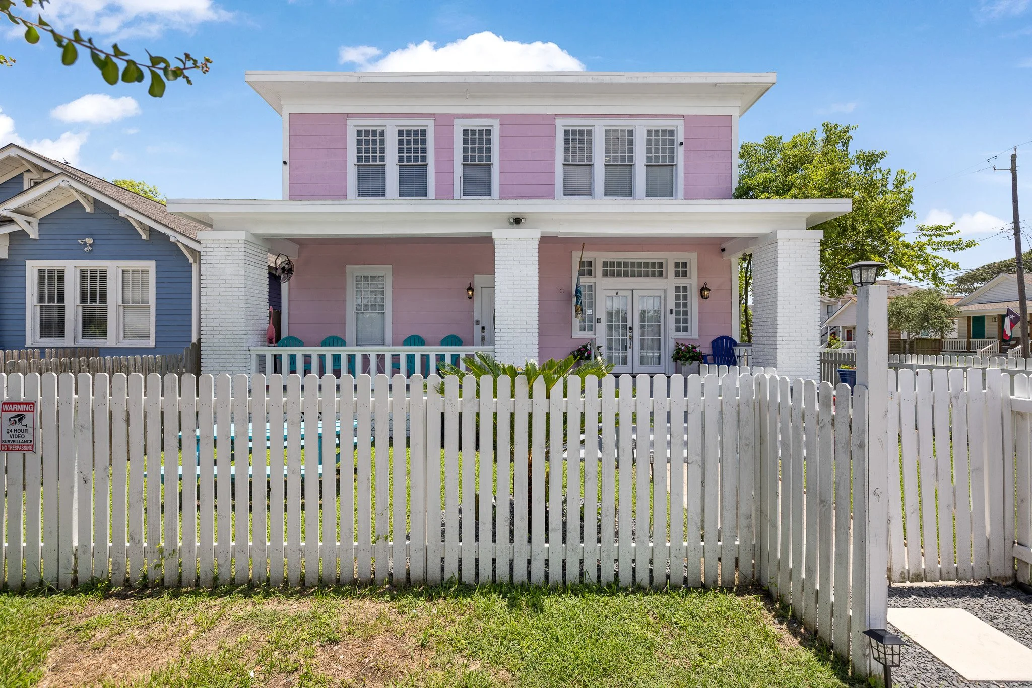 A colorful two-story house with pink and blue exterior, white porch and picket fence, located in a residential neighborhood on a sunny day.