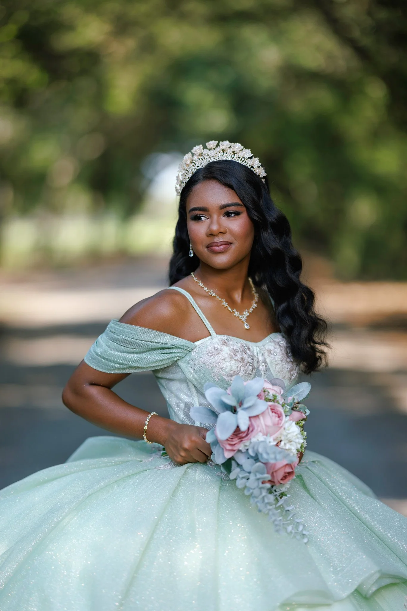 A woman dressed in a light green, off-the-shoulder gown with a puffy skirt, holding a bouquet of pink and white roses with greenery. She has dark, wavy hair, wears a pearl and diamond necklace, matching earrings, and a floral tiara, standing outdoors