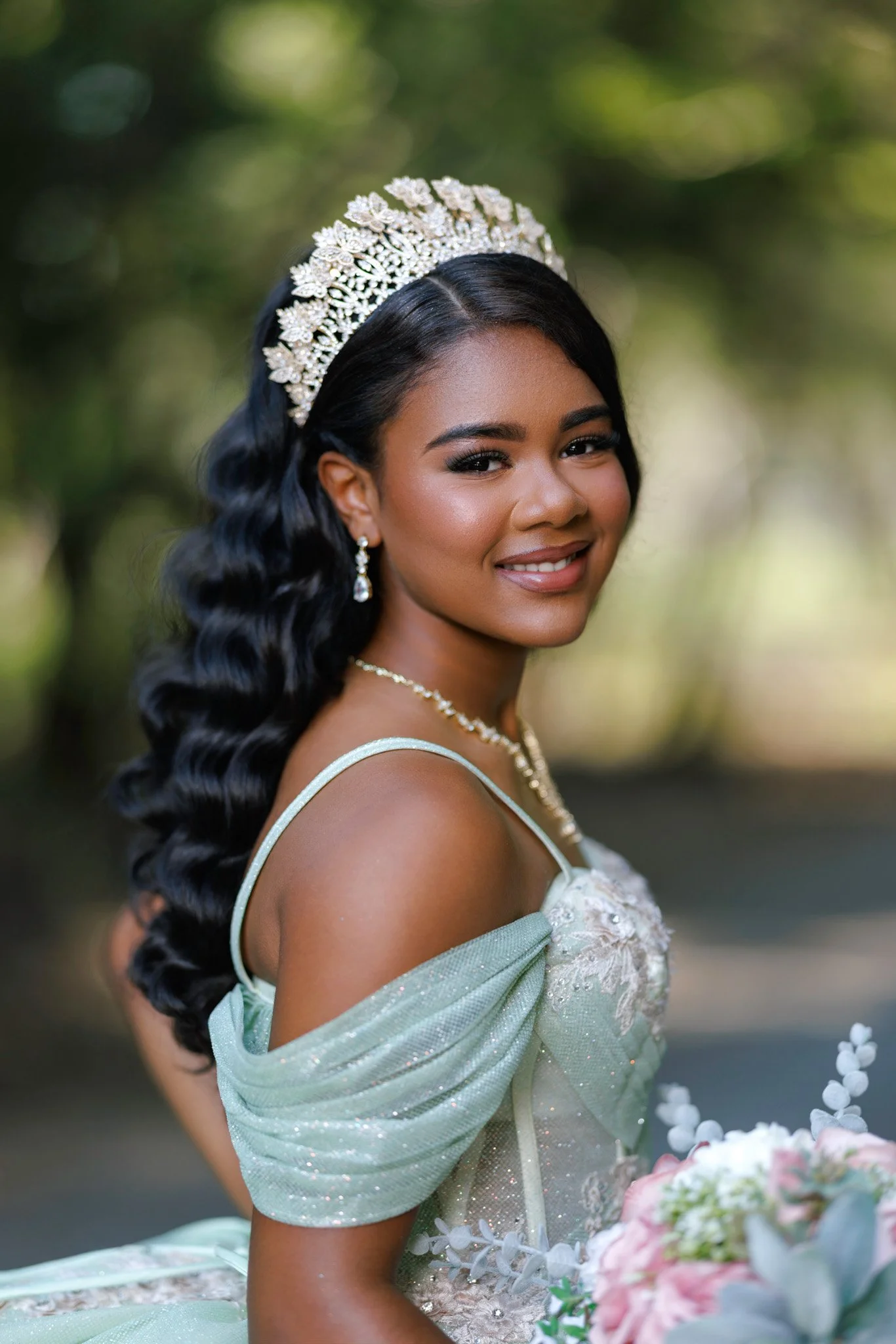 A bride with dark wavy hair wearing a sparkling tiara and pearl earrings, smiling outdoors with green blurred background, holding a bouquet of pink and white flowers.